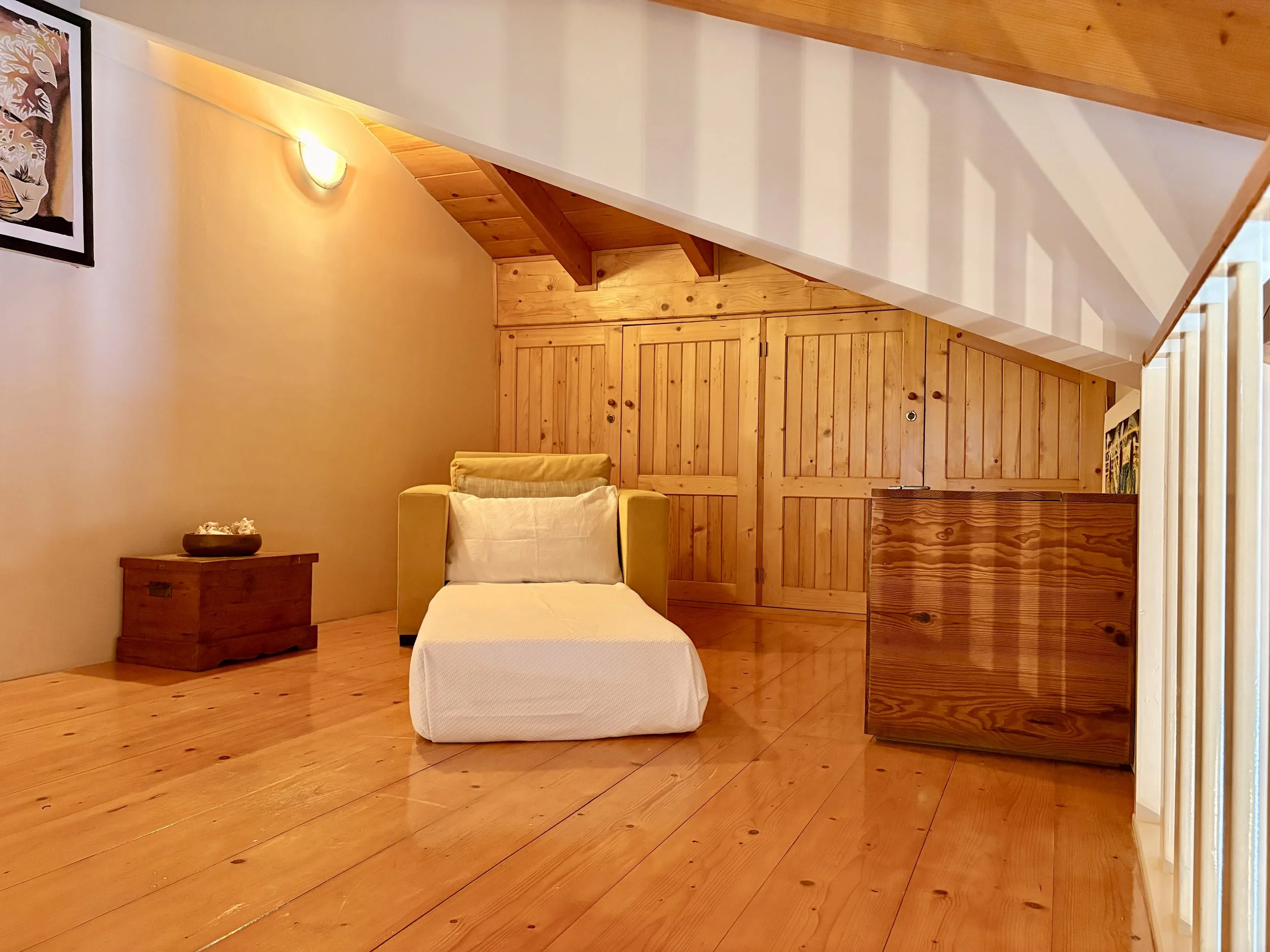 A cozy interior room with wooden walls and ceiling, a yellow armchair with a pillow, a side table with a bowl of shells, and a framed artwork on the wall.