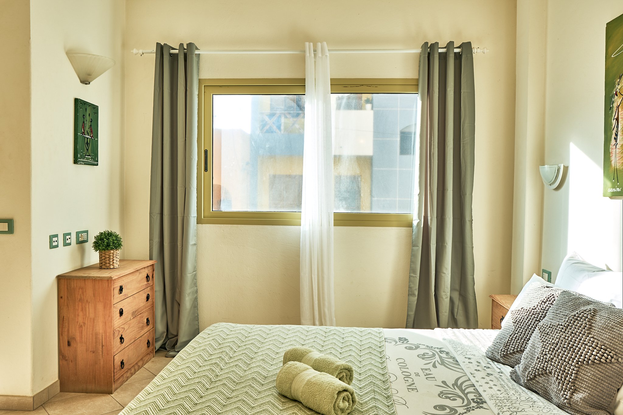 A bedroom with a window behind a white curtain, two small wooden dressers on either side, a bed with a patterned bedspread, rolled towels, and pillows, with artwork on the walls and a plant on one dresser.