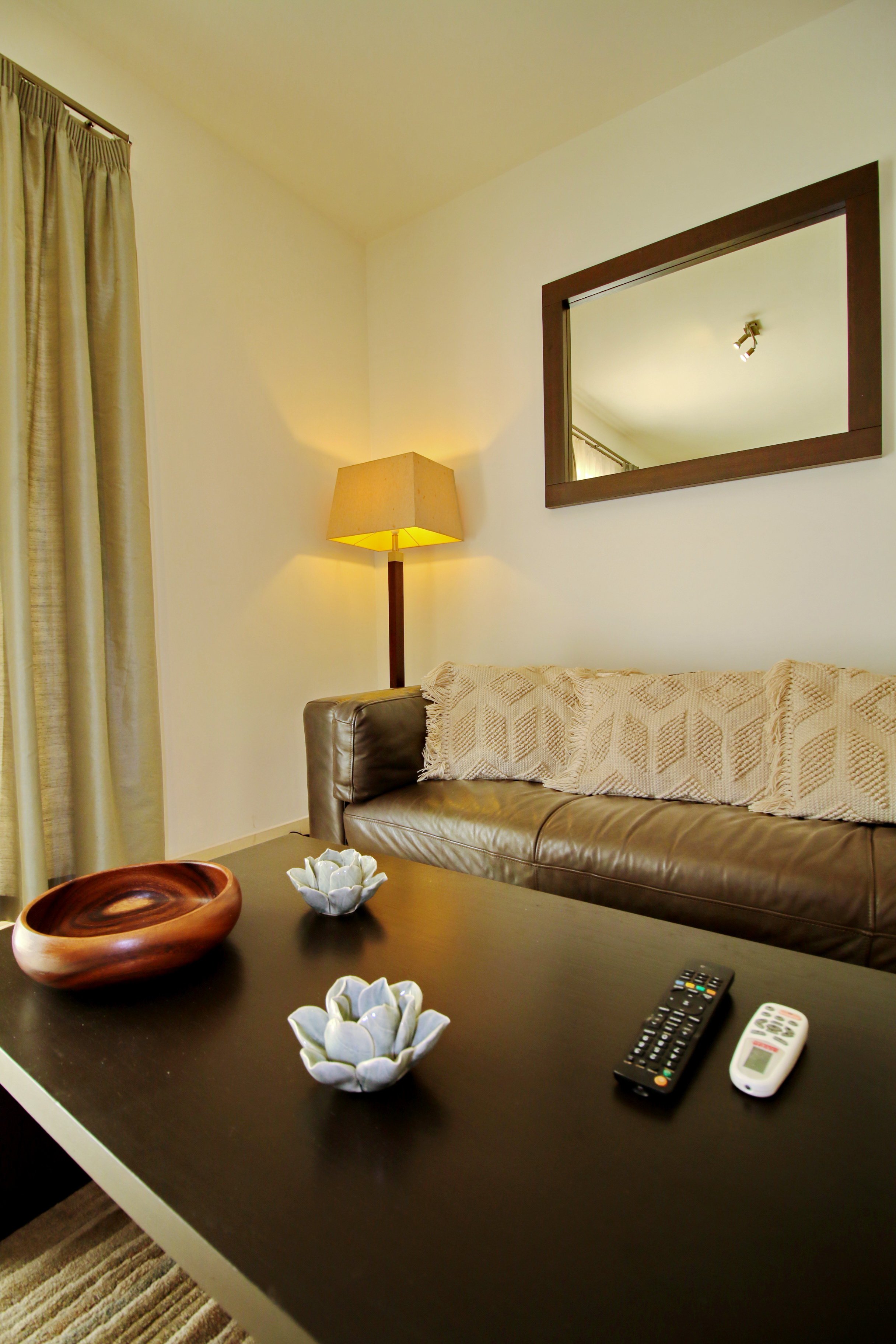 Living room with beige curtains, a lamp on a wood stand, a mirror on the wall, a beige couch with a textured blanket, a coffee table with decorative flower-shaped items, a wooden bowl, and remote controls.