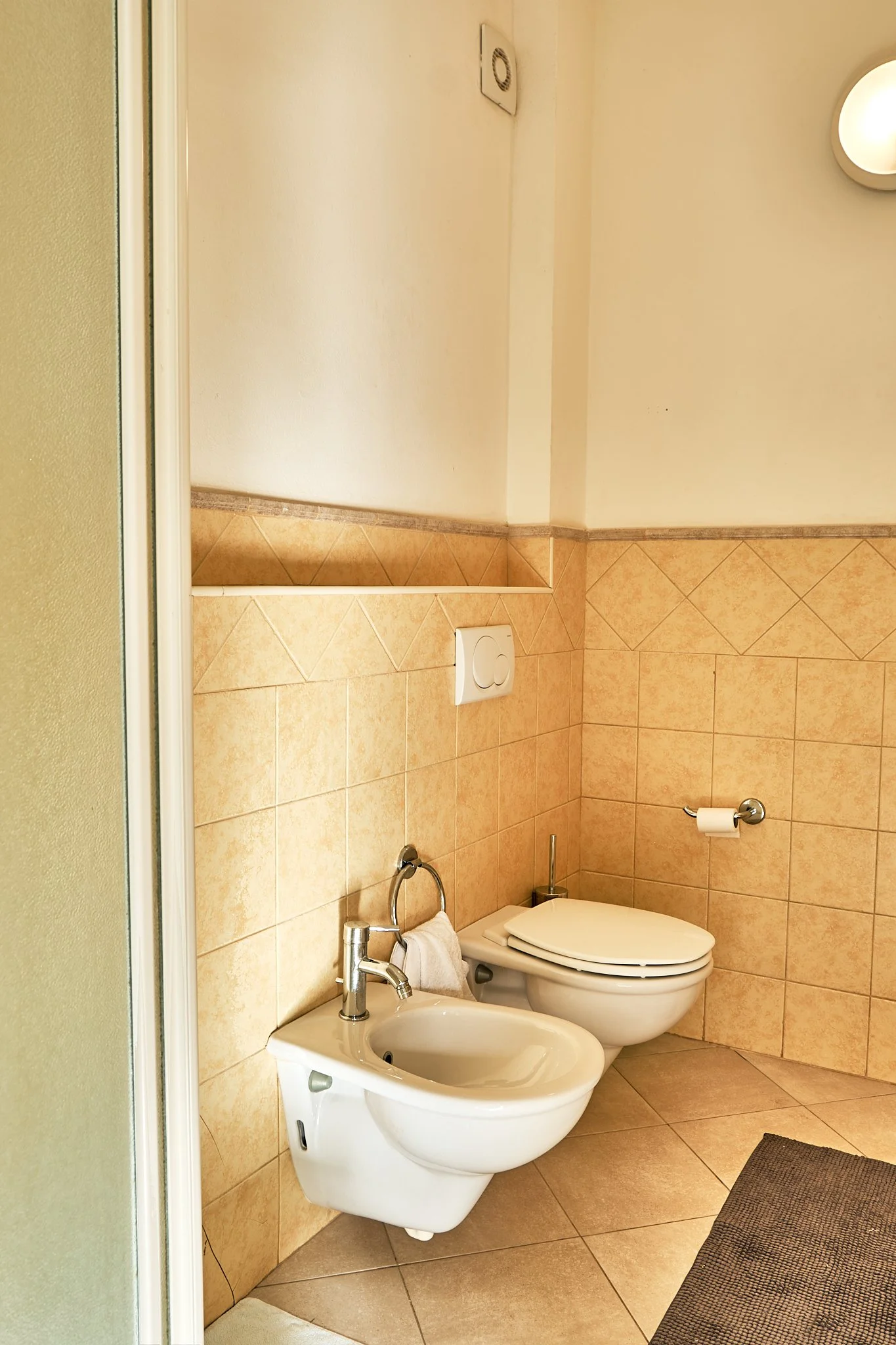 Bathroom with beige tiled walls and floor, toilet, bidet, toilet paper holder, towel holder, and ceiling vent.