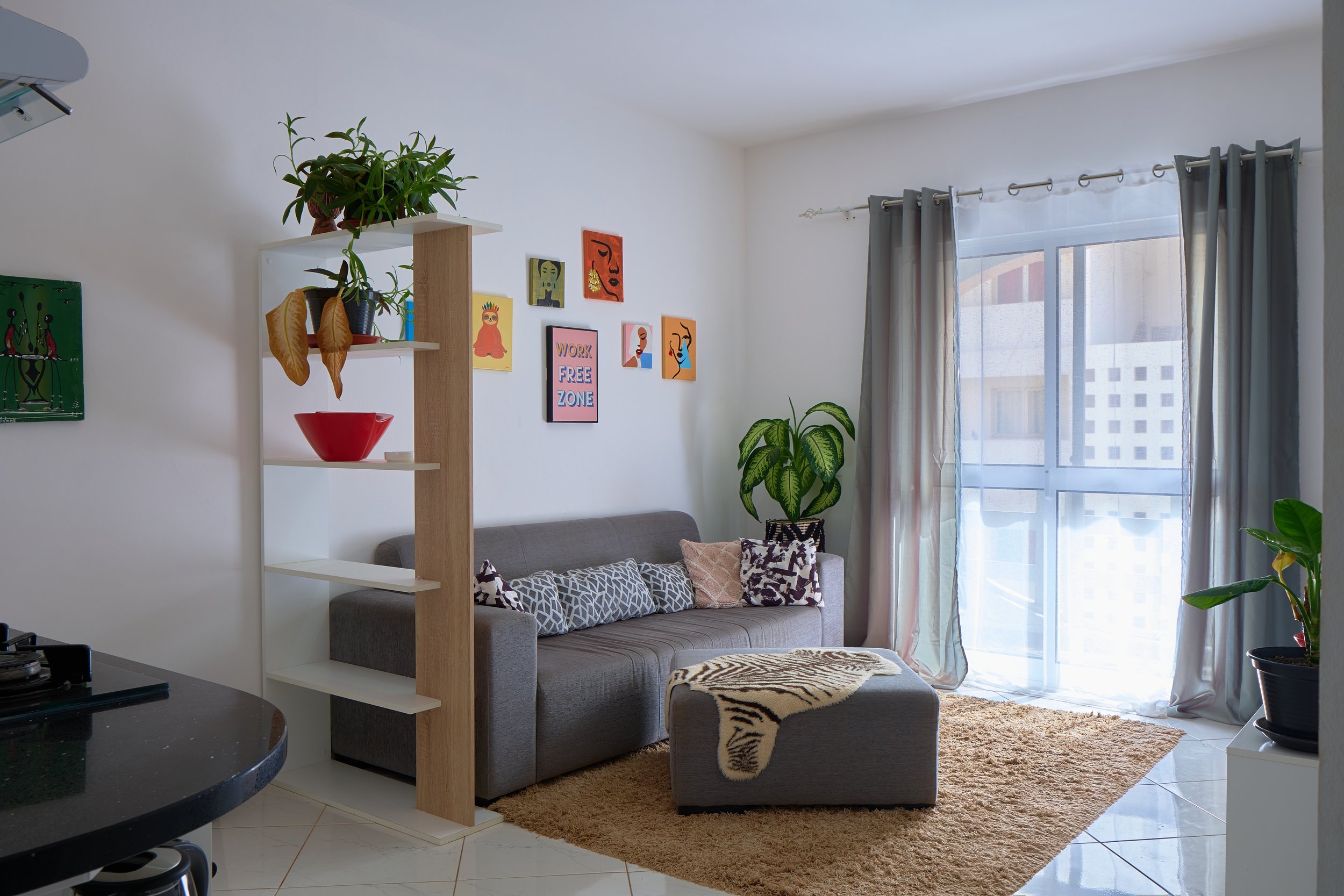 Living room with a gray sofa, patterned pillows, a beige rug with a zebra print, a large window with curtains, wall art, and indoor plants.
