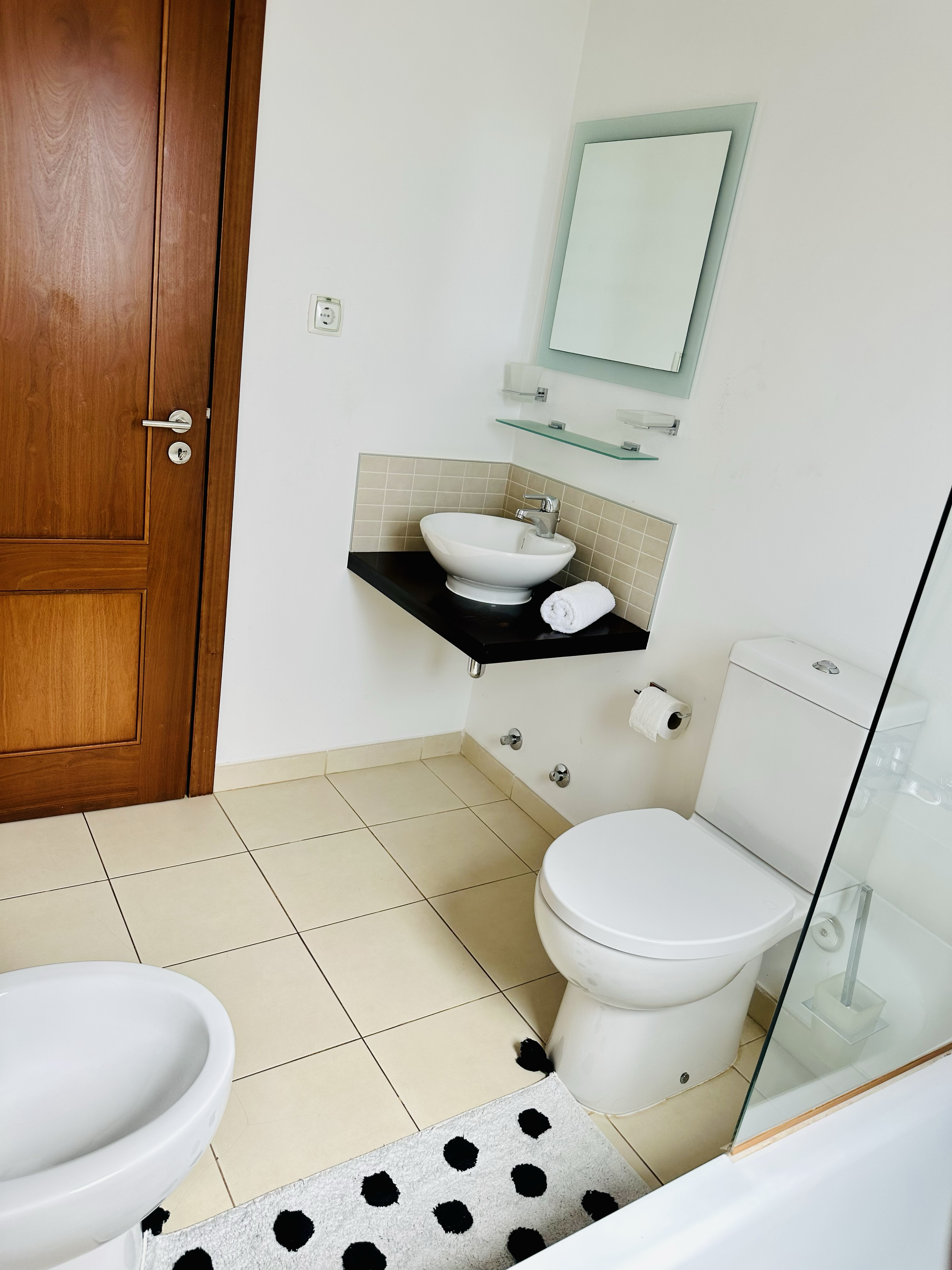 A modern bathroom featuring a wooden door, a white toilet, a small white basin with a chrome faucet, a mirror with a light frame, a glass shelf, beige tiles, and a black and white polka-dot rug.