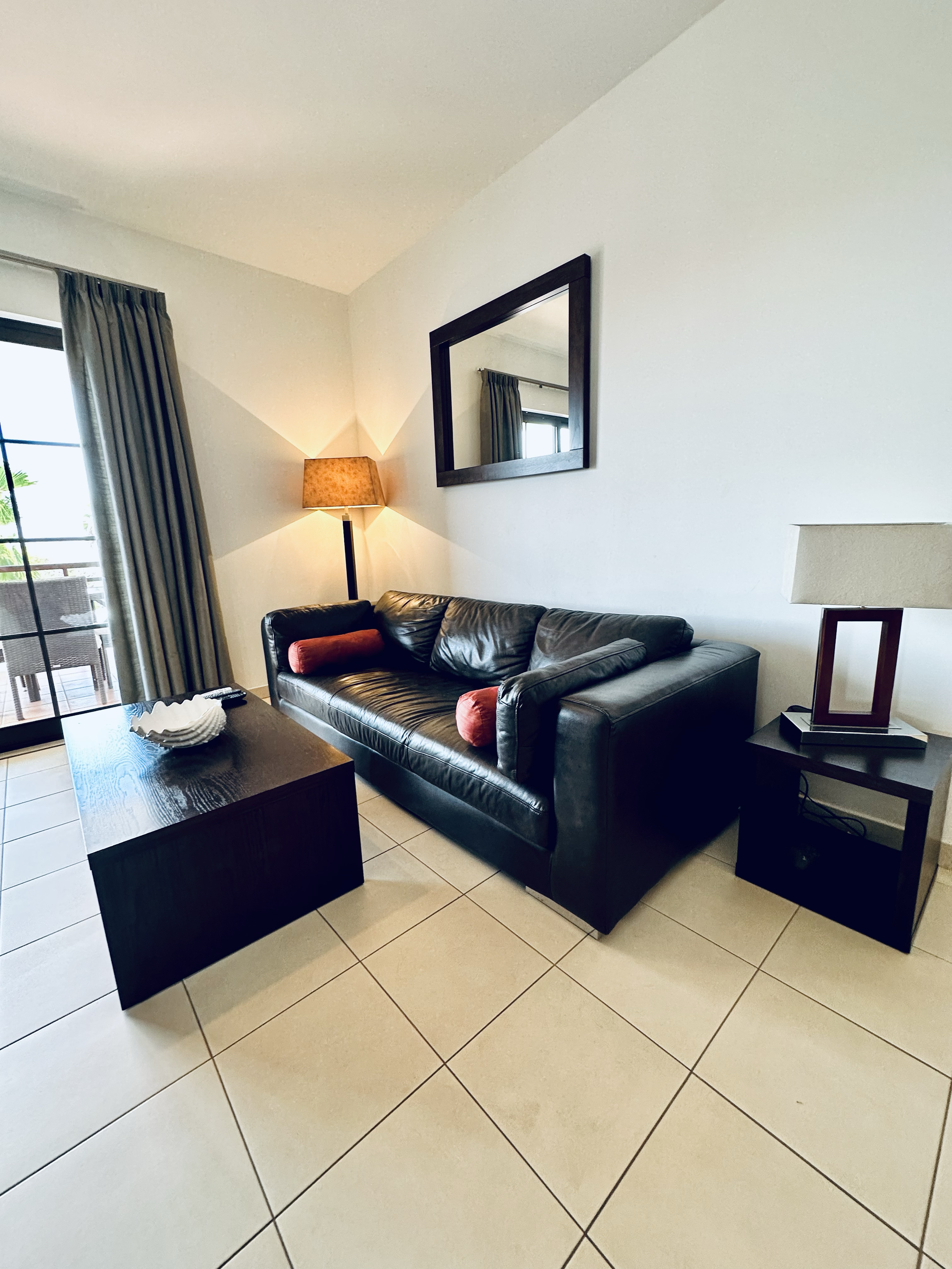Living room with a black leather couch, curtain-covered sliding glass door, side table with lamp, wall mirror, and tiled floor.