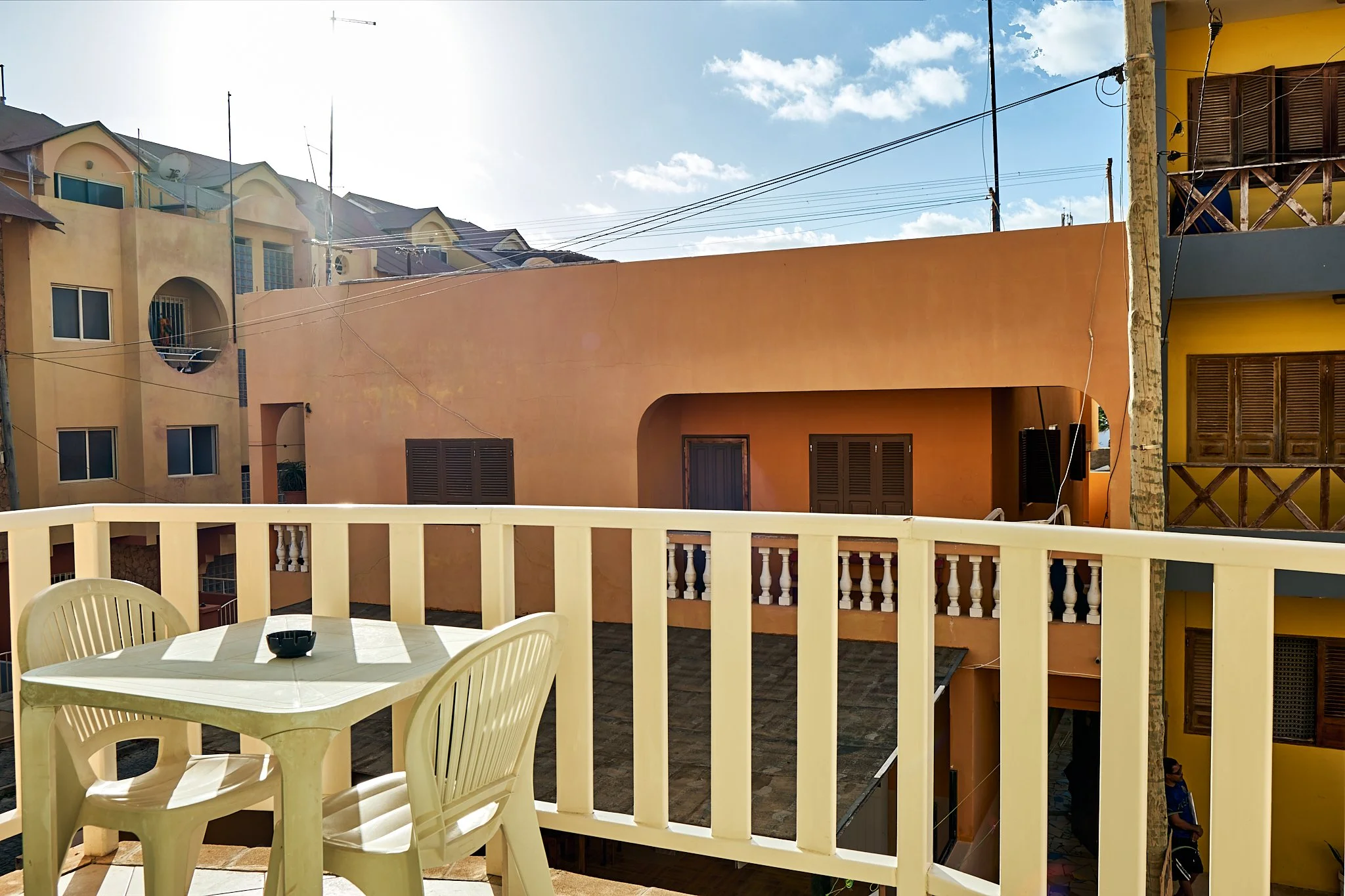 View from balcony with white plastic chairs and table overlooking neighboring multi-story colorful buildings under partly cloudy sky.