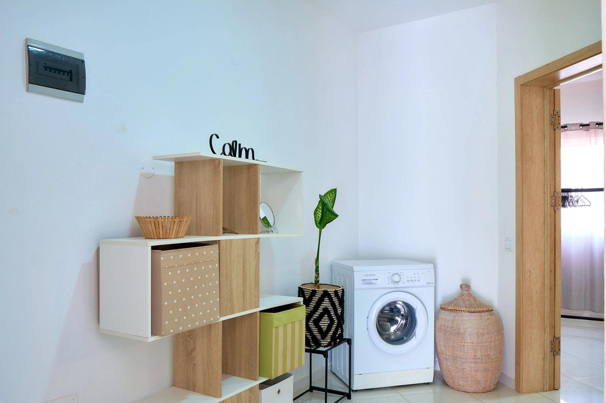 A laundry room with white walls and a closet door. There is a white washing machine labeled 'Telefac' in the corner, a wicker laundry basket, a tall woven basket, and a colorful striped storage bin. A wooden shelf unit holds various decor items, incl