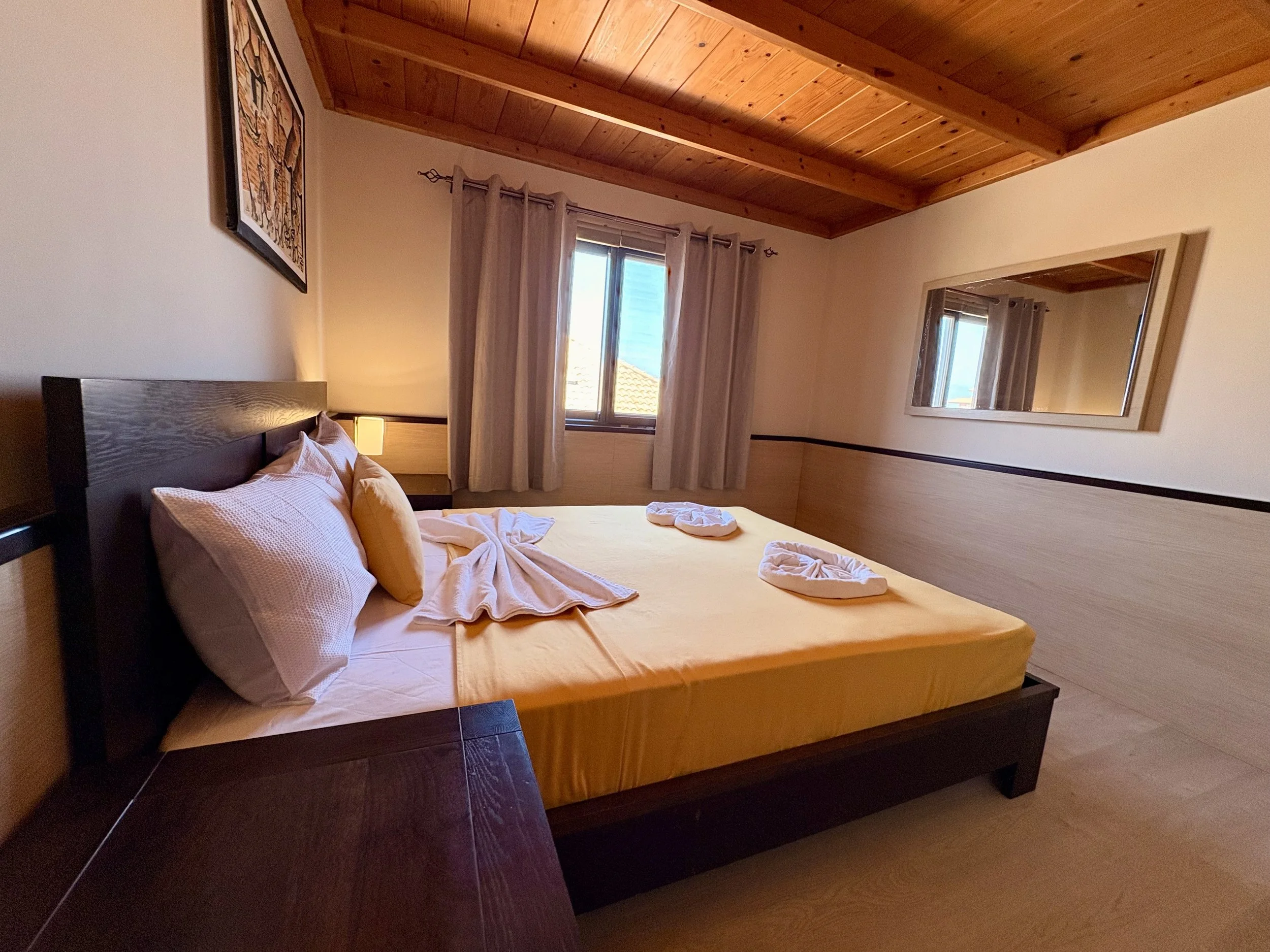 A bedroom with a wooden ceiling, a bed with white bedding and pillows, a window with pink curtains, a large mirror on the wall, and a piece of artwork hanging above the bed.