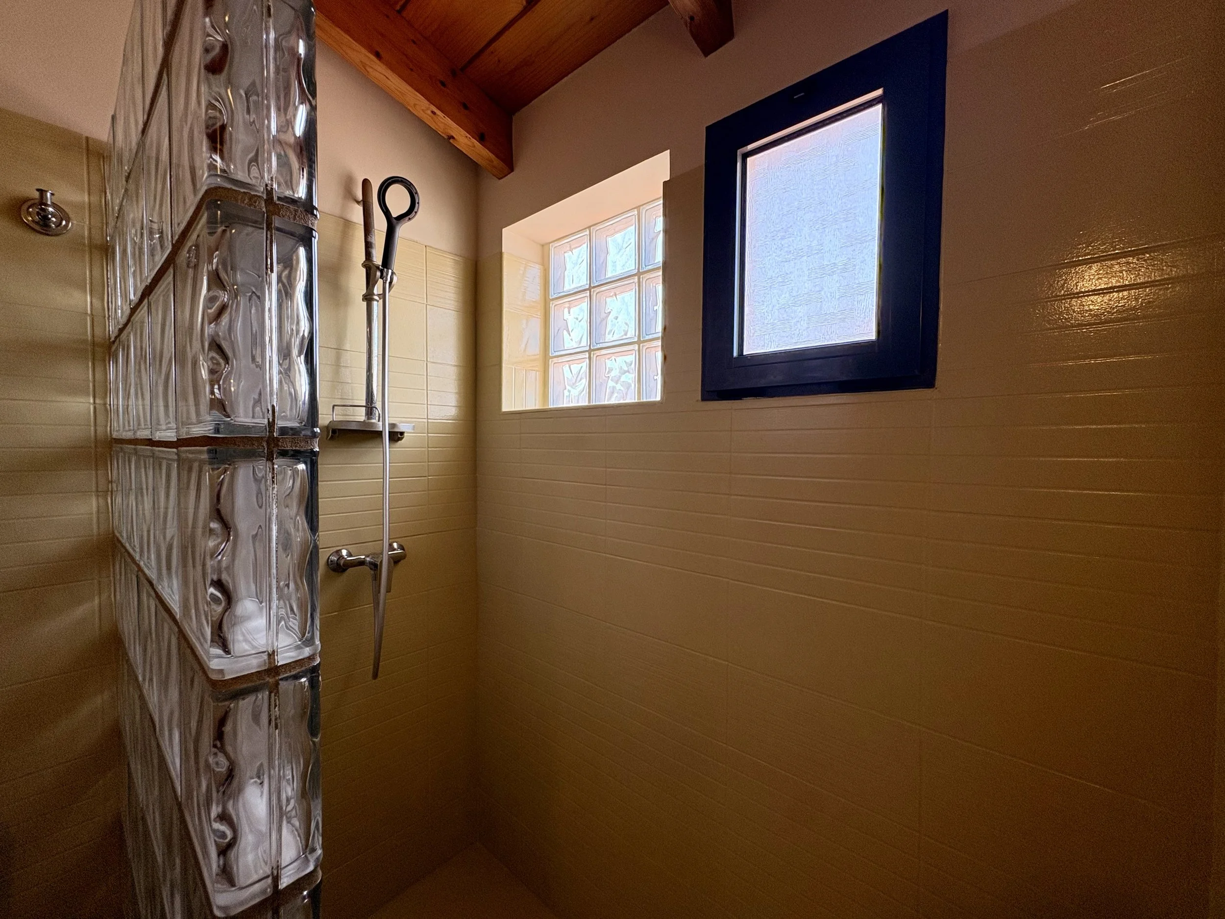 A bathroom shower with wood ceiling, beige tiles, a glass block window, dark blue framed window, and a chrome shelf with glass bottles