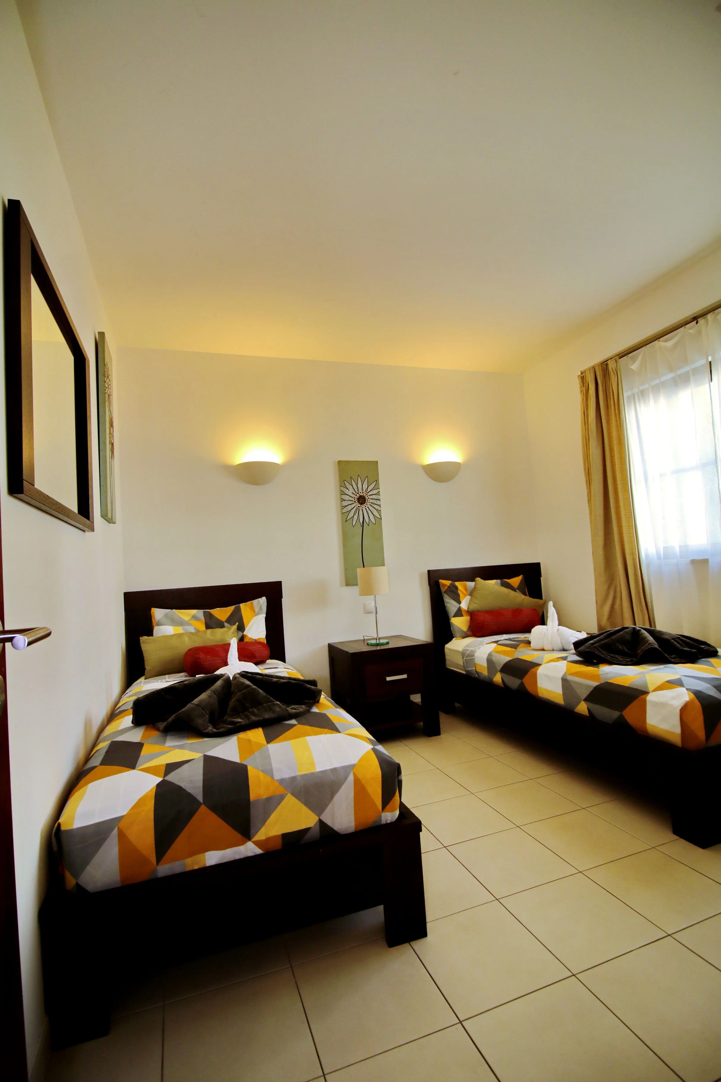 A hotel room with two twin beds, each with geometric patterned bedding in yellow, gray, white, and black. The beds have pillows, including red, yellow, and beige. A nightstand with a lamp is between the beds, with a framed wall art featuring a flower