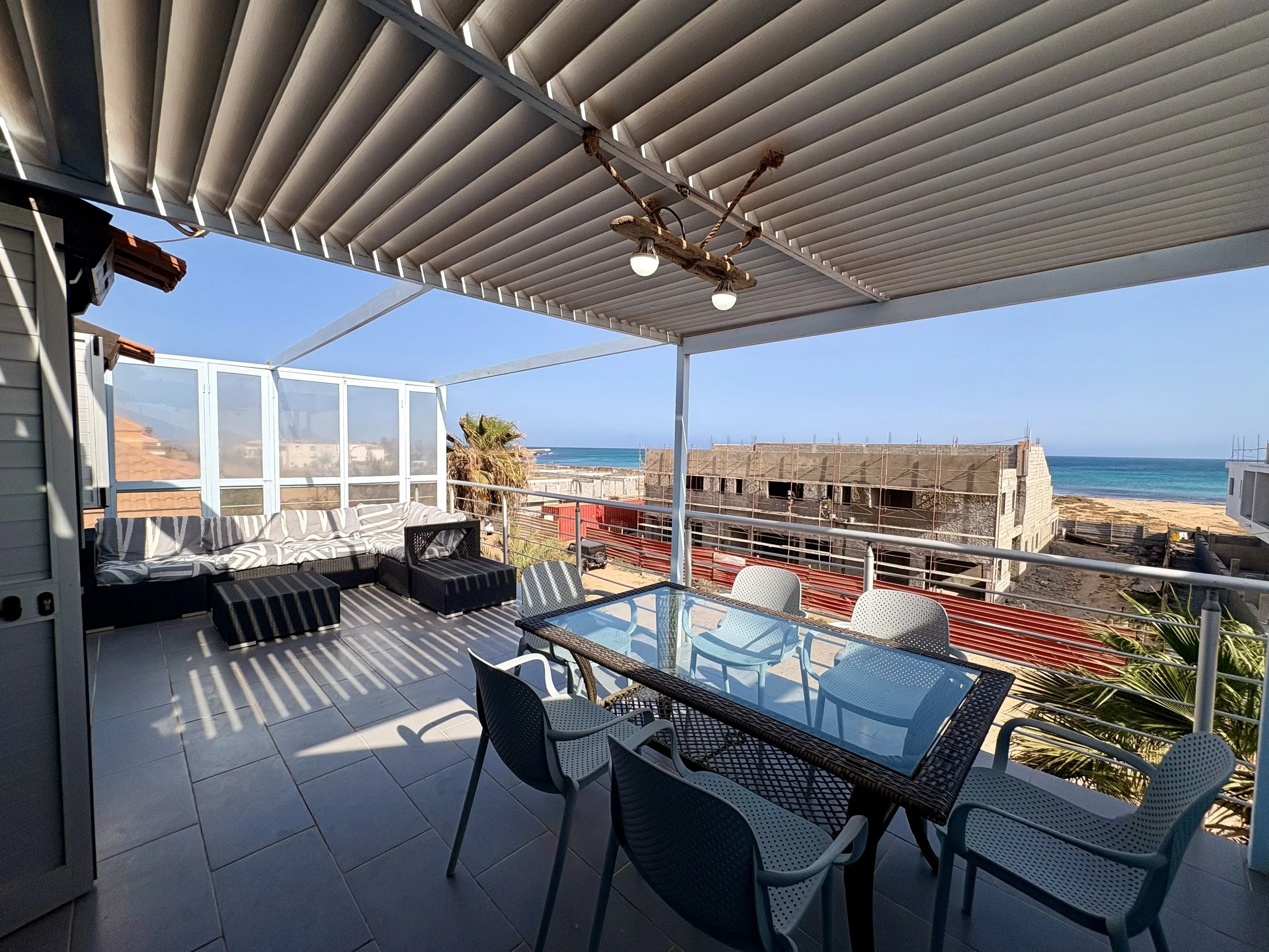 Balcony with outdoor seating overlooking a beach and ocean, with a blue sky. Furniture includes a glass-topped table and white plastic chairs, with a cushioned outdoor sofa. Overhead is a metal and wood chandelier, with a partially covered ceiling.