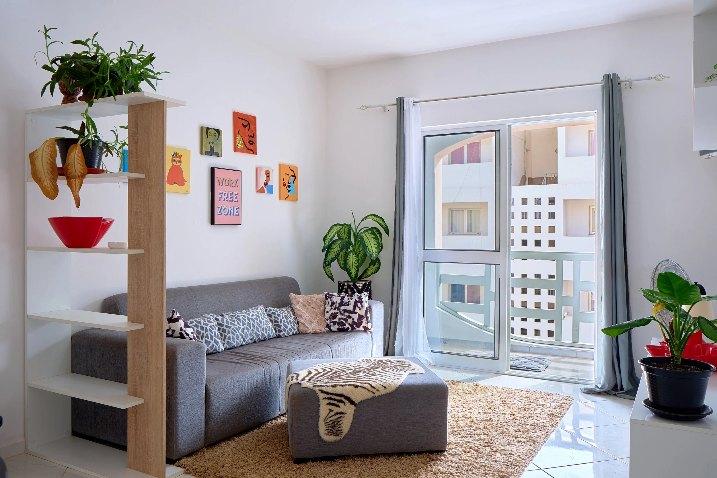 Living room with gray sofa, decorative pillows, and a zebra-print blanket, near large open window with white and gray curtains, potted plants, and wall art.