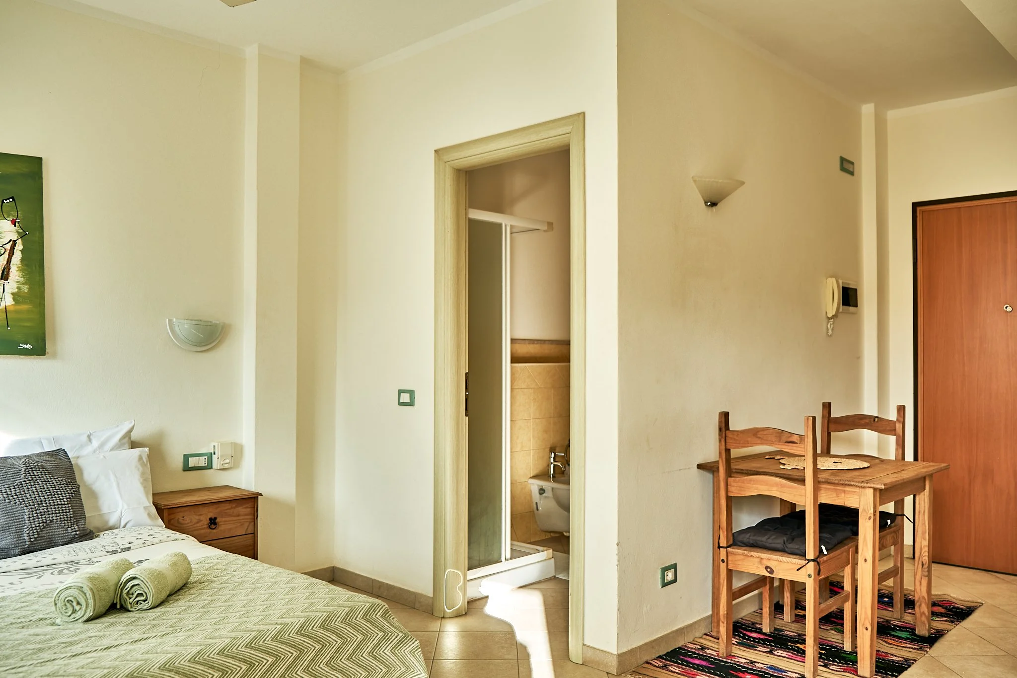 A bedroom view showing part of a bed with rolled towels, a wooden nightstand, and a wall-mounted light fixture. In the background, an open door leads to a bathroom with tiles, a toilet, and a sink. The room has cream-colored walls, a wooden dining ta