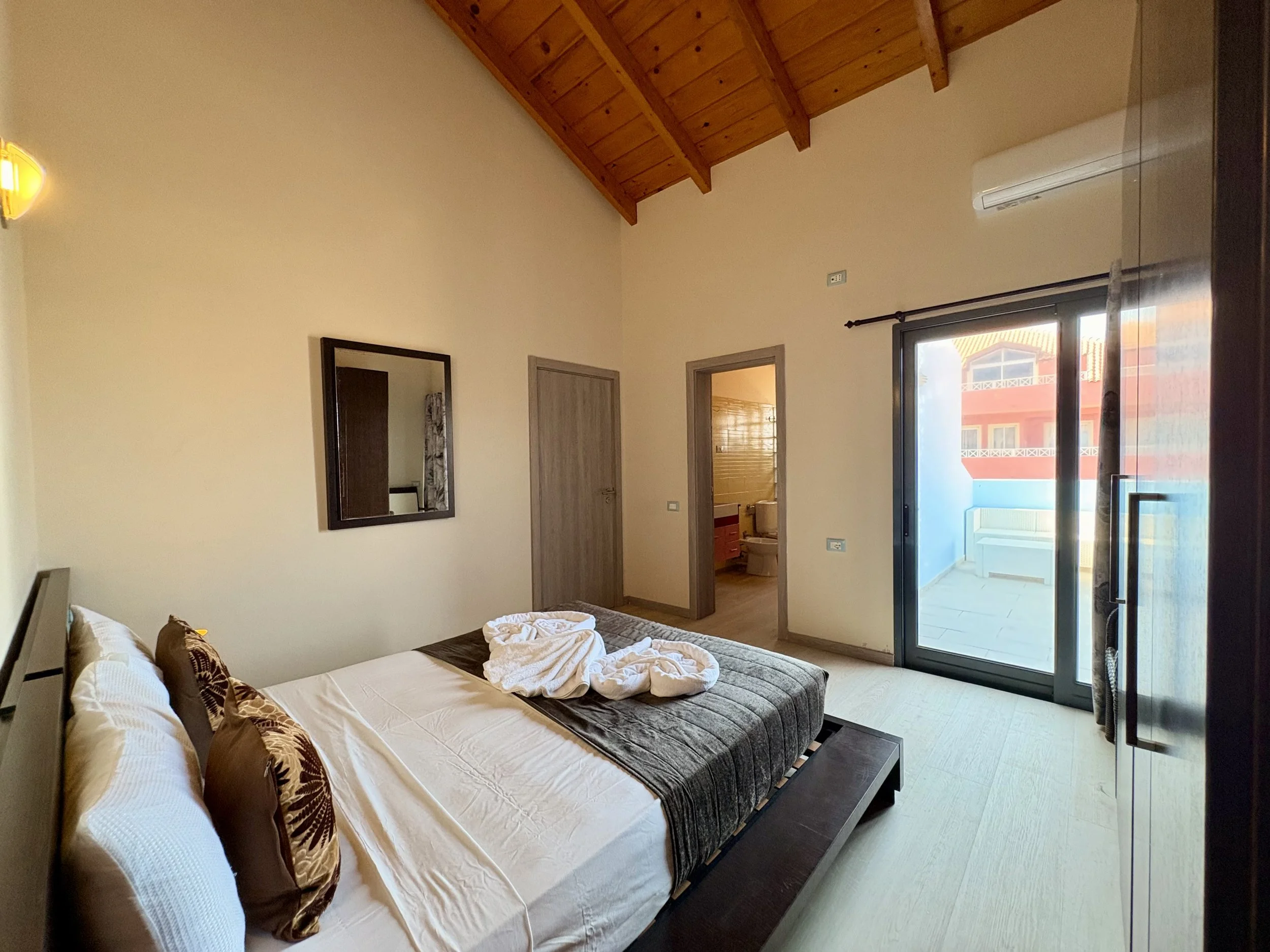 A bedroom with a bed, tossed bedding, and decorative pillows. The room has a high wooden ceiling, a wall mirror, an air conditioning unit, and large sliding glass doors leading to a balcony or patio with a view of neighboring buildings.