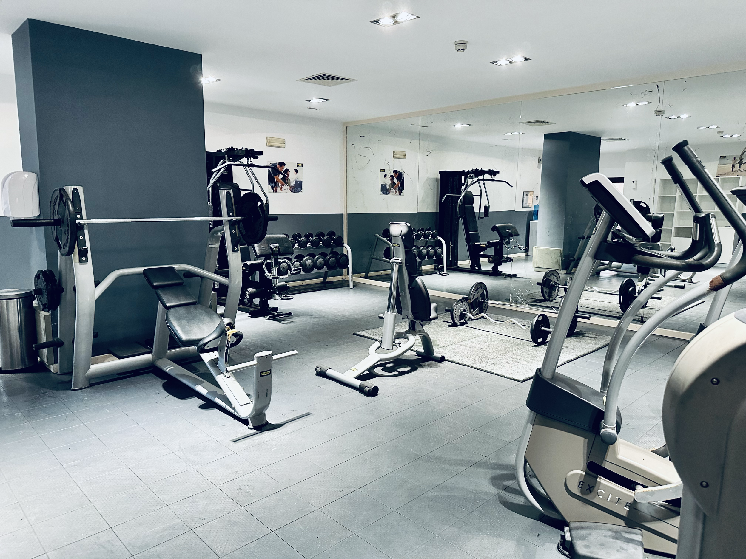 A gym with various exercise equipment including weight machines, dumbbells, and cardio machines, with a large mirror on one wall reflecting the equipment and some people working out in the background.