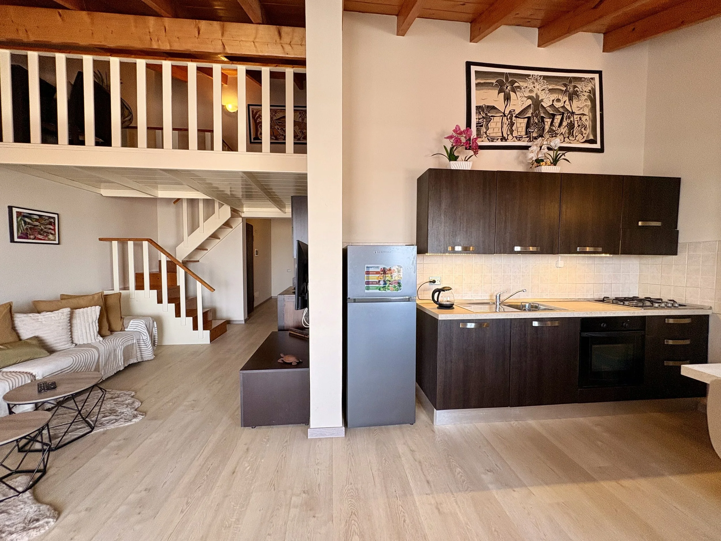 Open-concept living room and kitchen with wooden ceiling beams, framed artwork on the wall, potted plants, a sofa with striped cushions, and a small coffee table.