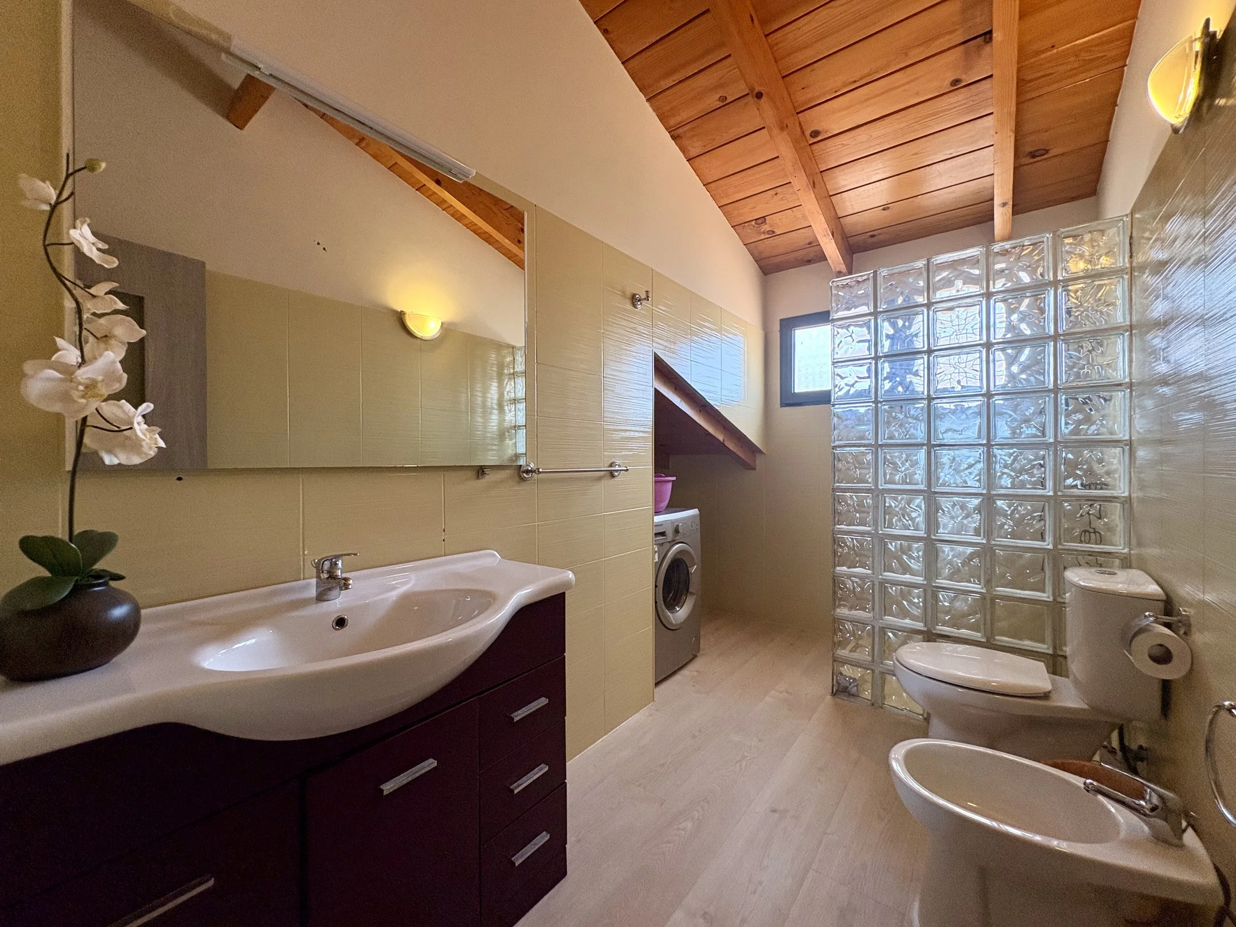 A spacious bathroom with a curved white sink, a dark cabinet with drawers, beige tiles, and a washing machine. There are two toilets, a bidet, and a glass block wall. The ceiling is wooden with beams, and there are small windows and wall lights.