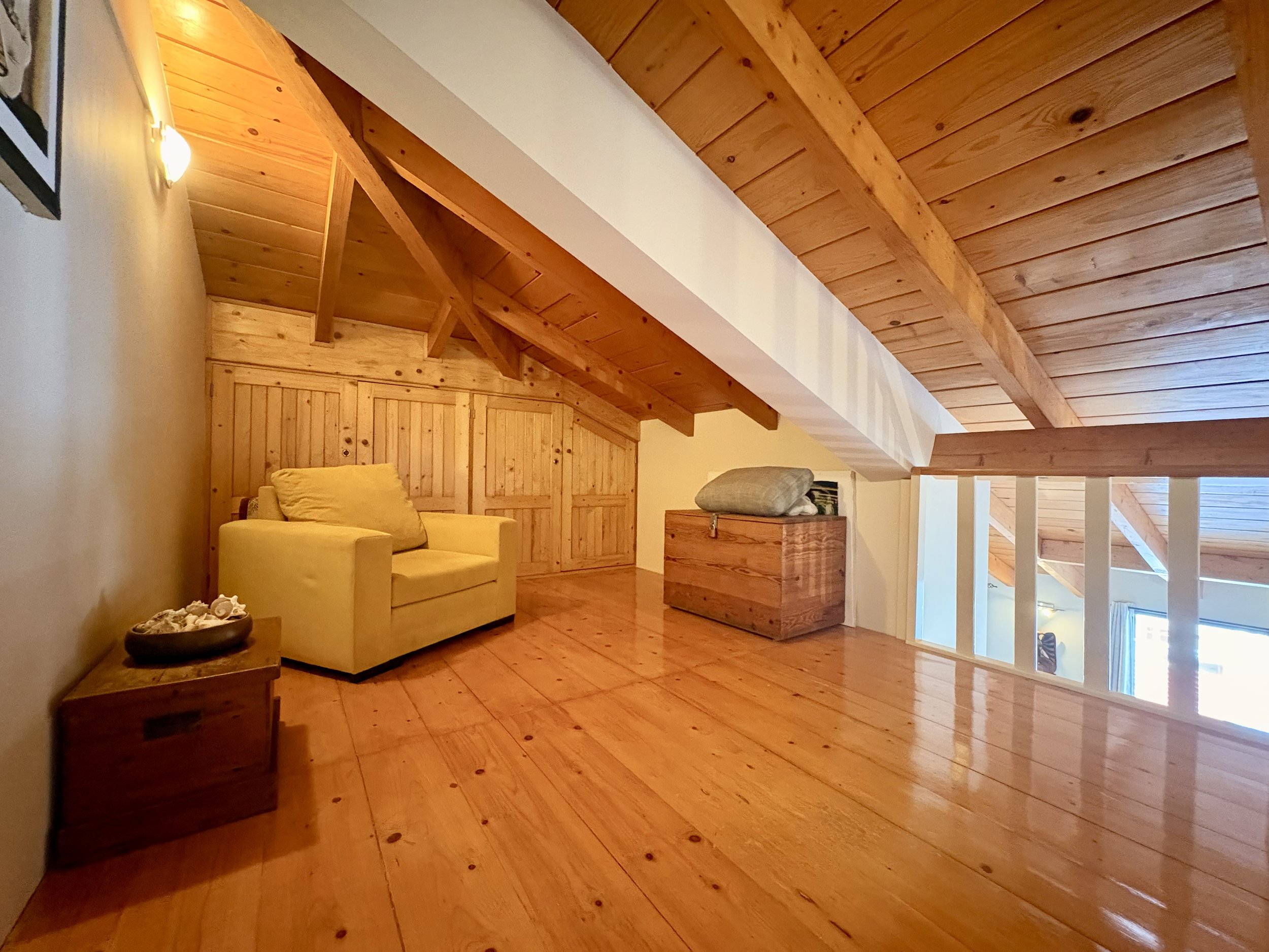 View of a cozy attic living space with wooden paneling on the ceiling and walls, a yellow armchair, and a small side table with a shell decoration.