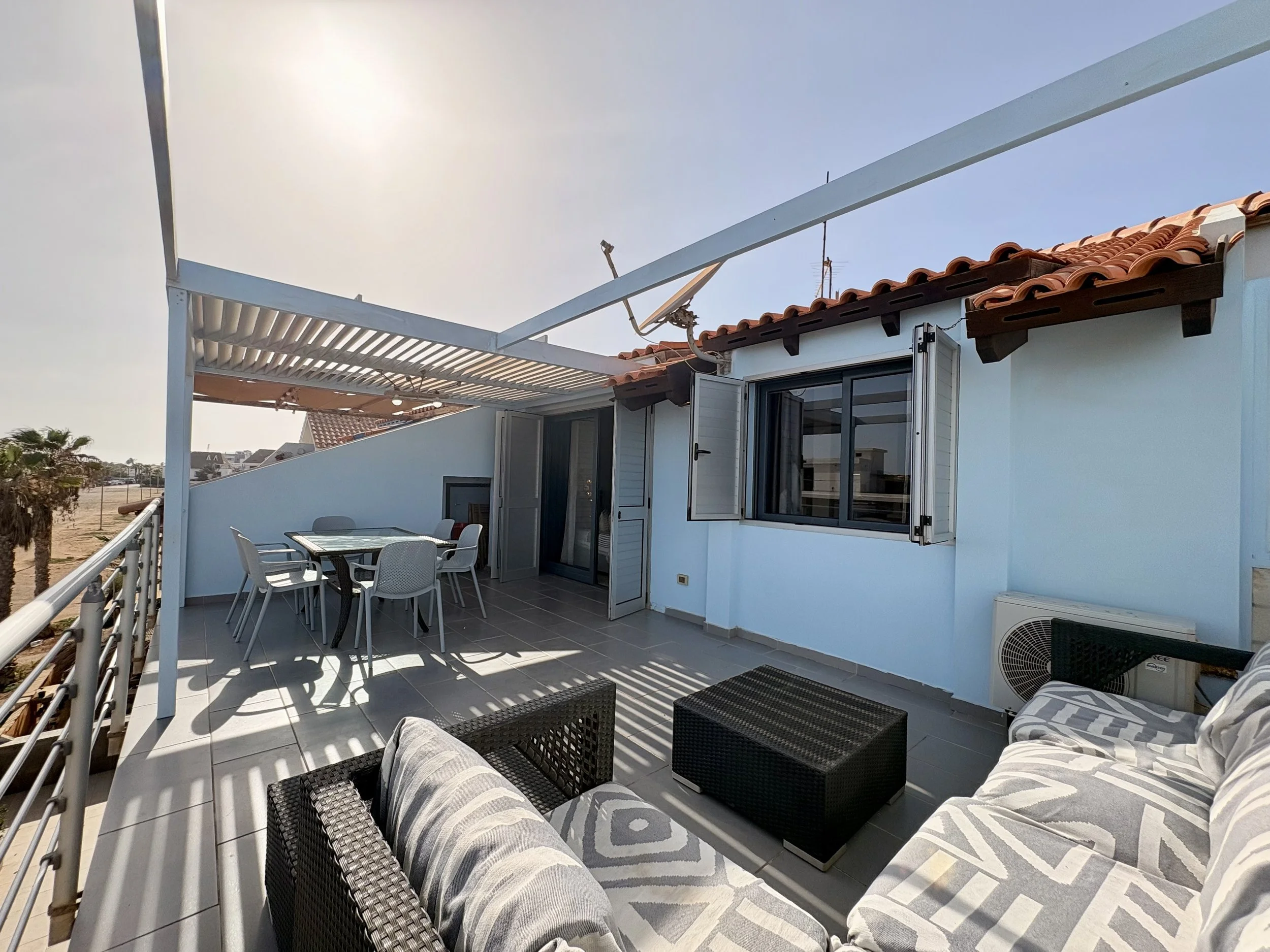 An outdoor balcony with patio furniture including white chairs and a glass table, black wicker seating with patterned cushions, a shaded area with a slatted roof, and a view of neighboring houses with red tile roofs under a clear blue sky.