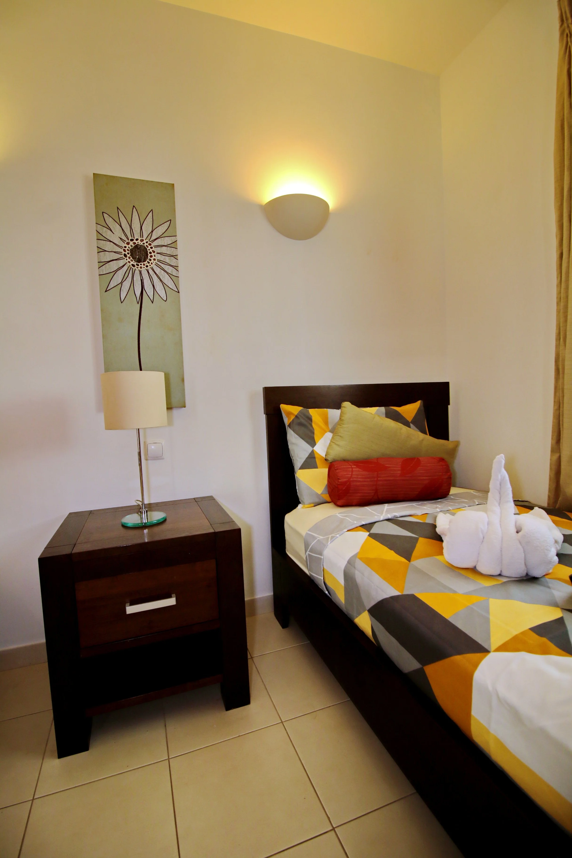 A bedroom featuring a bed with a geometric yellow, gray, and white patterned bedspread, several pillows including a yellow and gray one, and a towel folded into the shape of a swan on the bed. To the left, there is a dark wooden nightstand with a dra