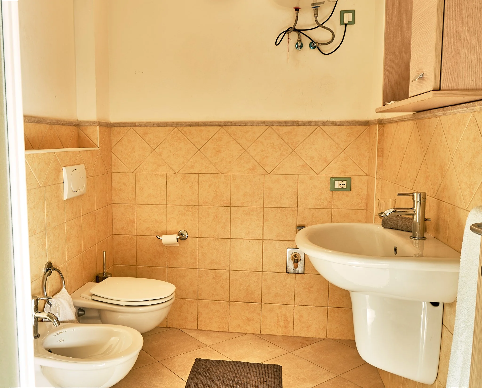 A small bathroom with a beige tile floor and walls. There is a white toilet with a bidet attachment, a sink with a silver faucet, and a mirror above the sink. A towel hangs on a bar next to the toilet. There are two electrical outlets on the wall, on