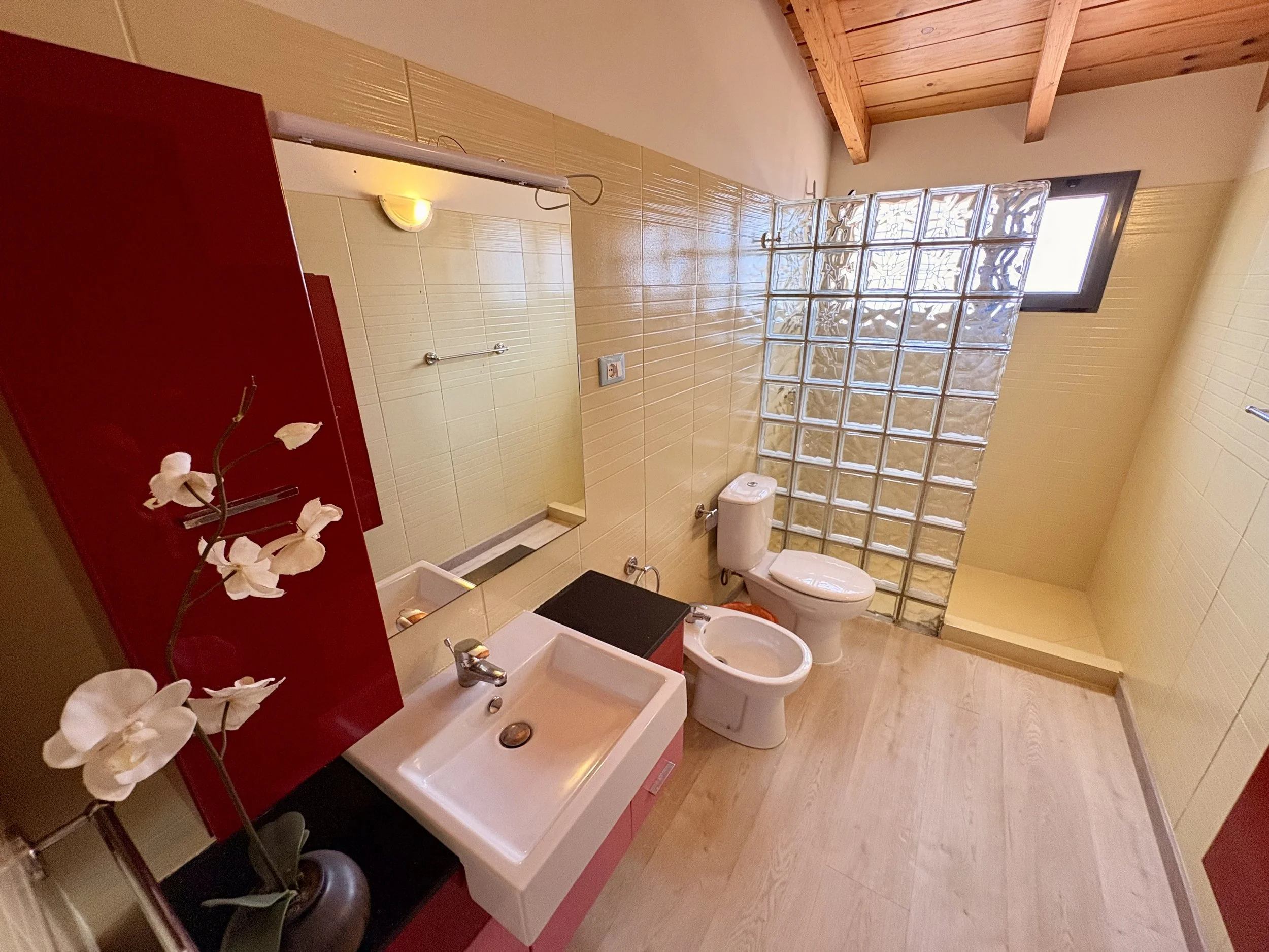 A bathroom with wooden ceiling, beige tiled walls, a small sink, a mirror, a bidet, a toilet, a window with glass blocks, and a red cabinet with a flower arrangement.
