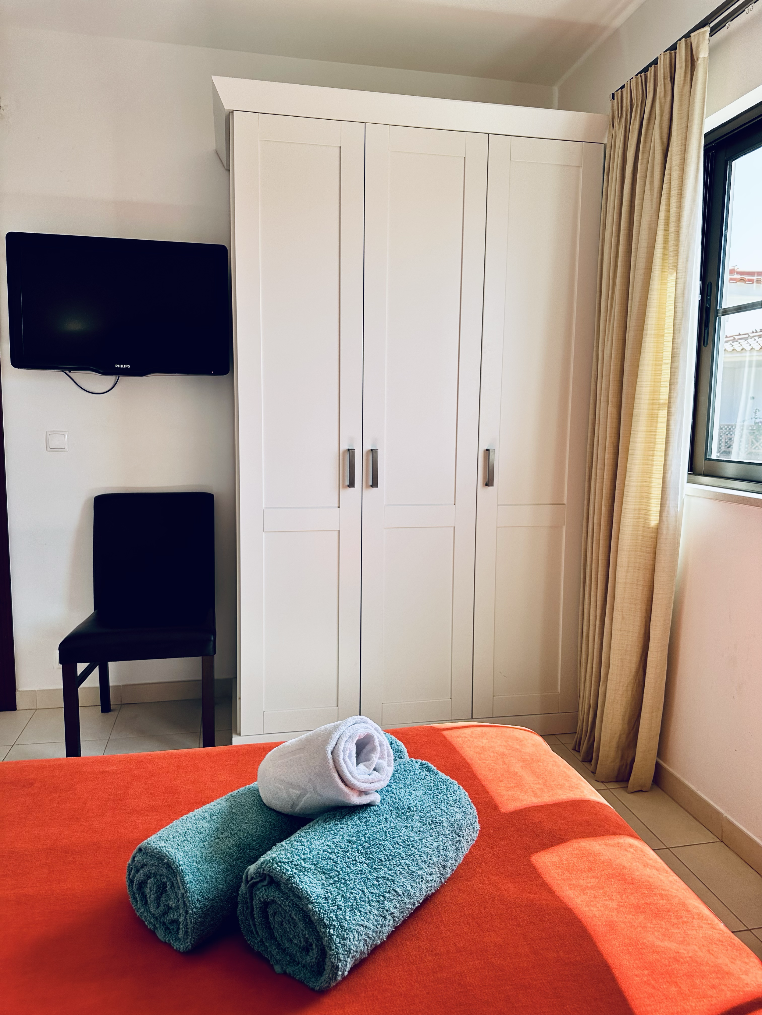 A bedroom with an orange bedspread, two rolled teal towels and a white towel shaped like a swan on the bed. A black chair, a white wardrobe, a window with beige curtains, and a wall-mounted flat-screen TV are visible.