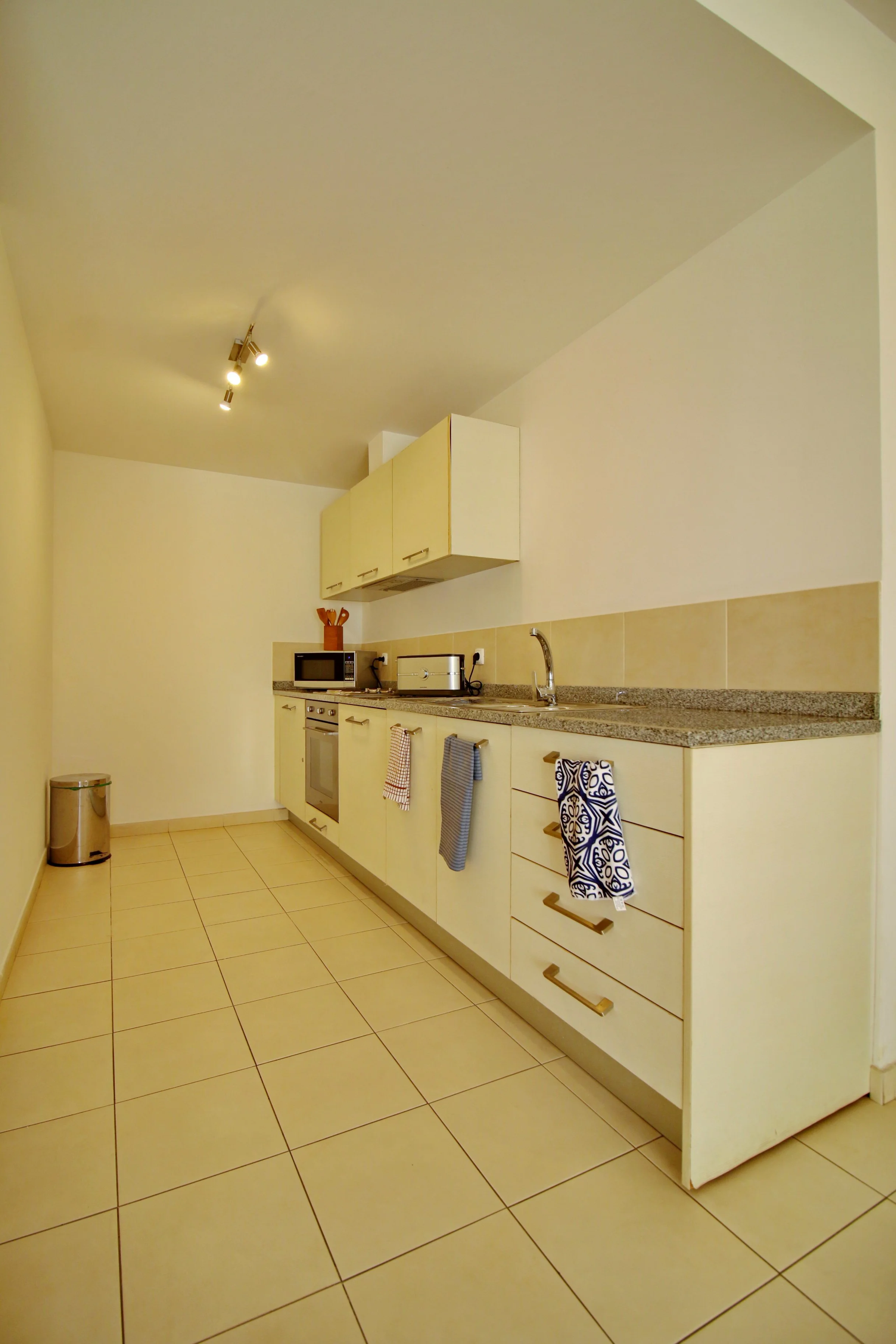 Kitchen with cream-colored cabinets, granite countertop, tiled floor, microwave, toaster, utensil holder, and towels hanging on cabinet handles.
