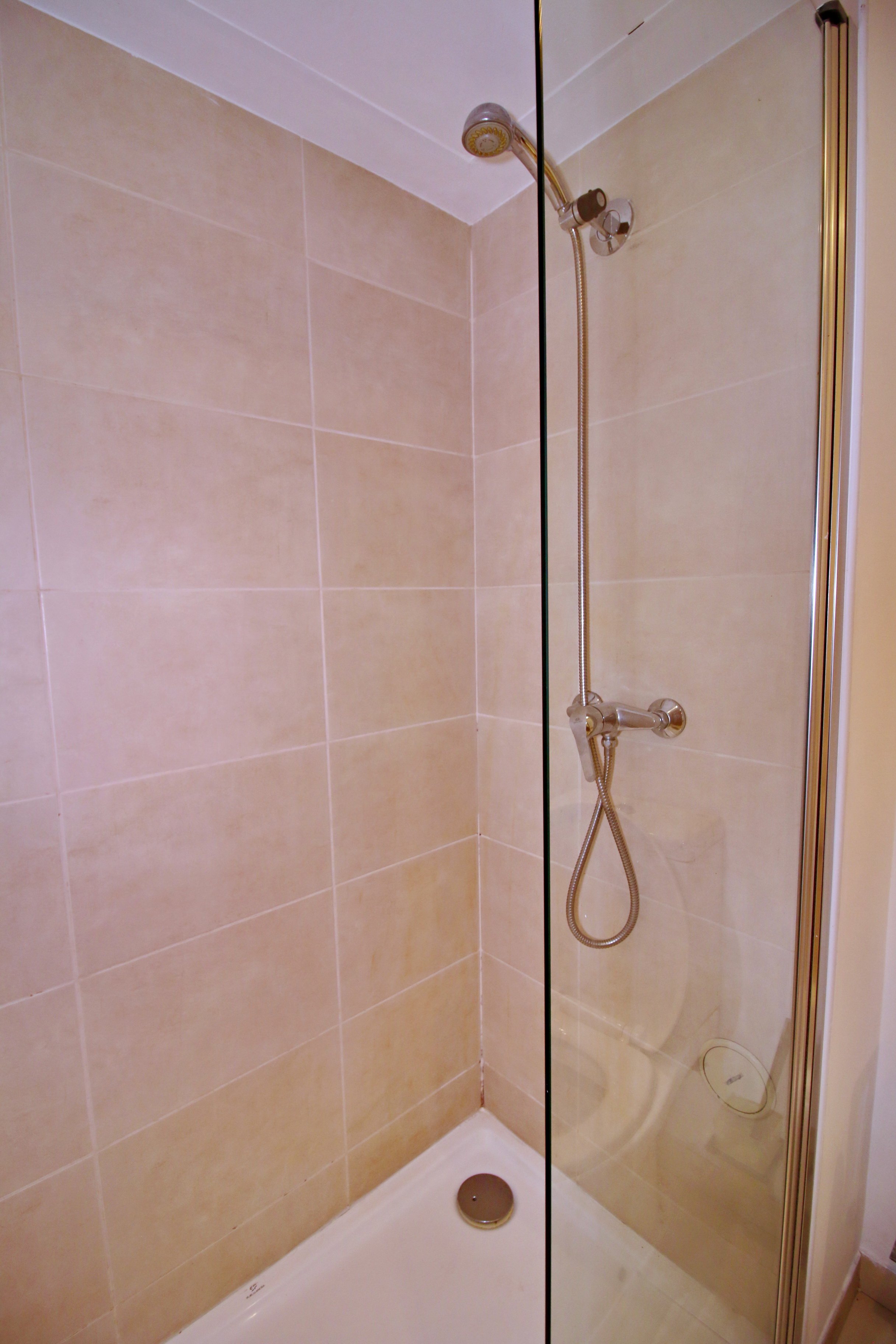 A beige tiled shower with a glass door, a wall-mounted handheld showerhead, and a drain on the shower floor.