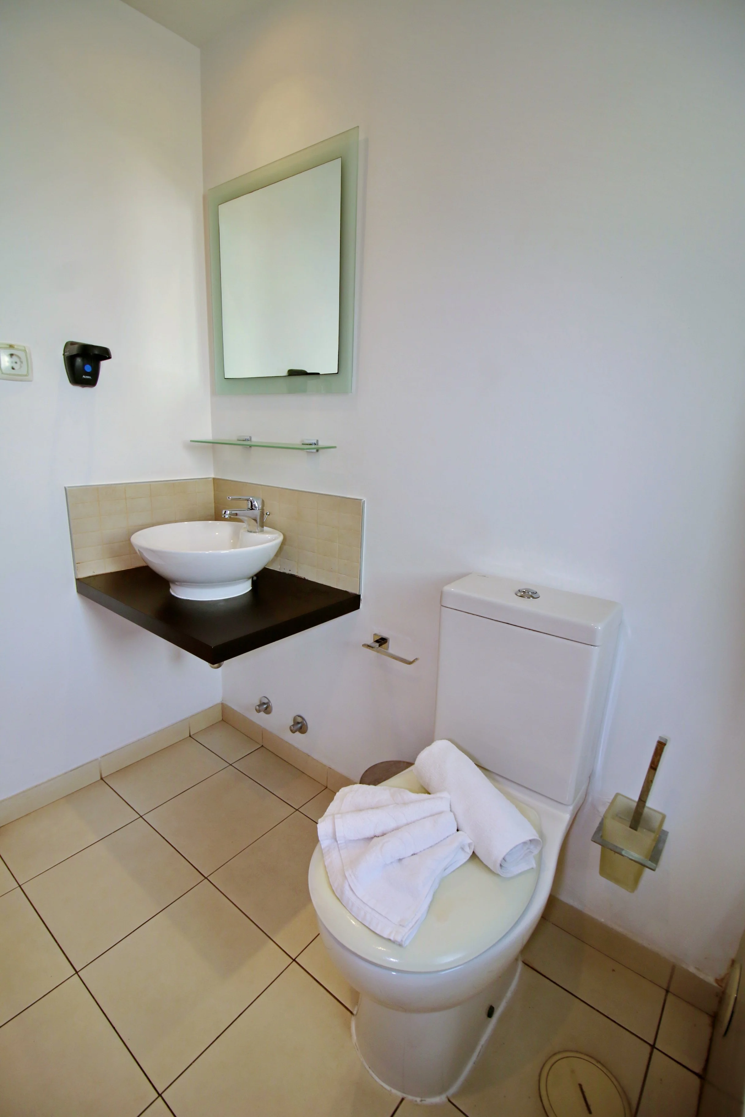 A bathroom with a white toilet, towels on the toilet lid, a sink with a black countertop, a mirror, a soap dispenser, and a drain on the beige tiled floor.