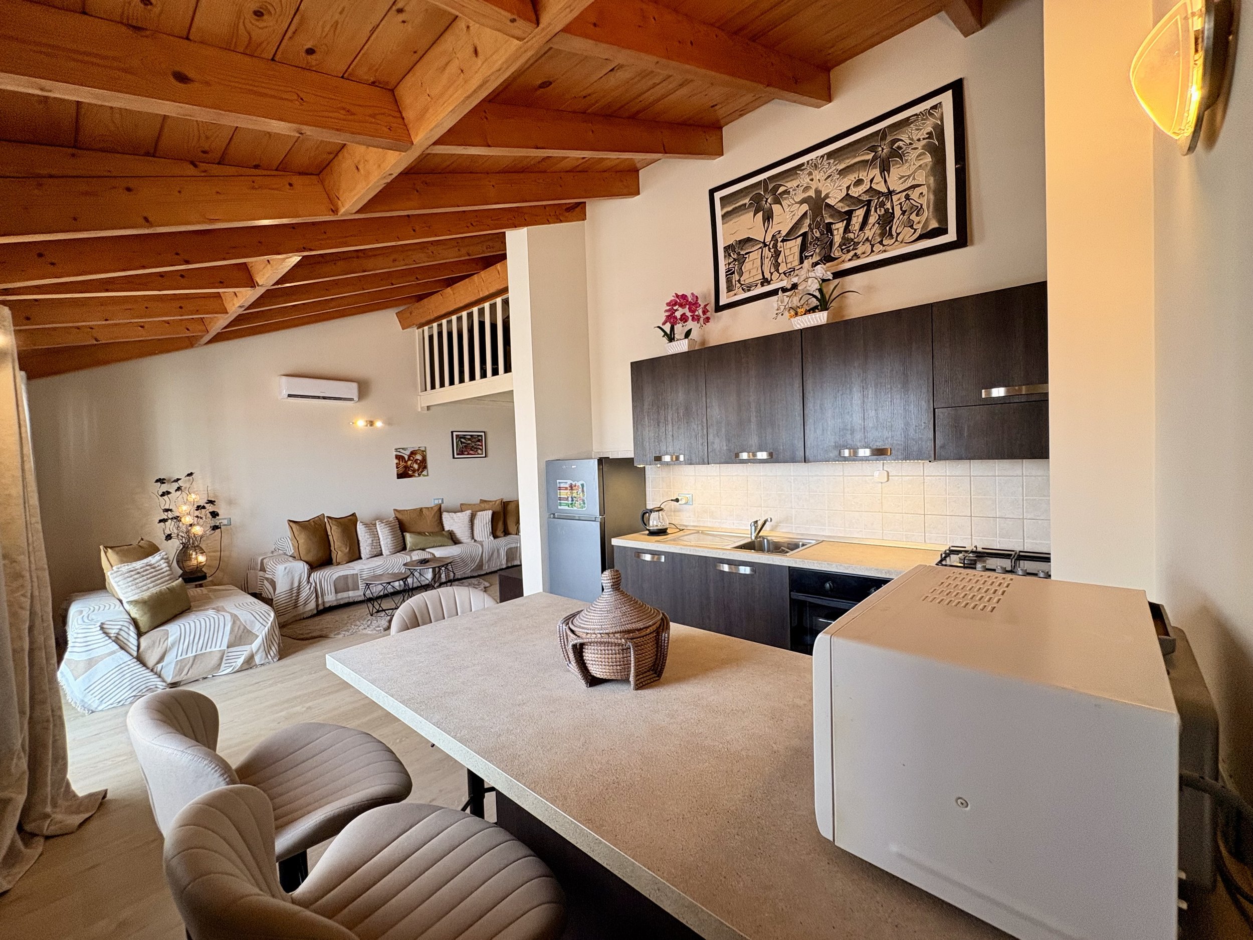 Living room with a wooden ceiling, beige upholstered chairs, a sofa, a painting on the wall, and a kitchen area with dark cabinets and a countertop.