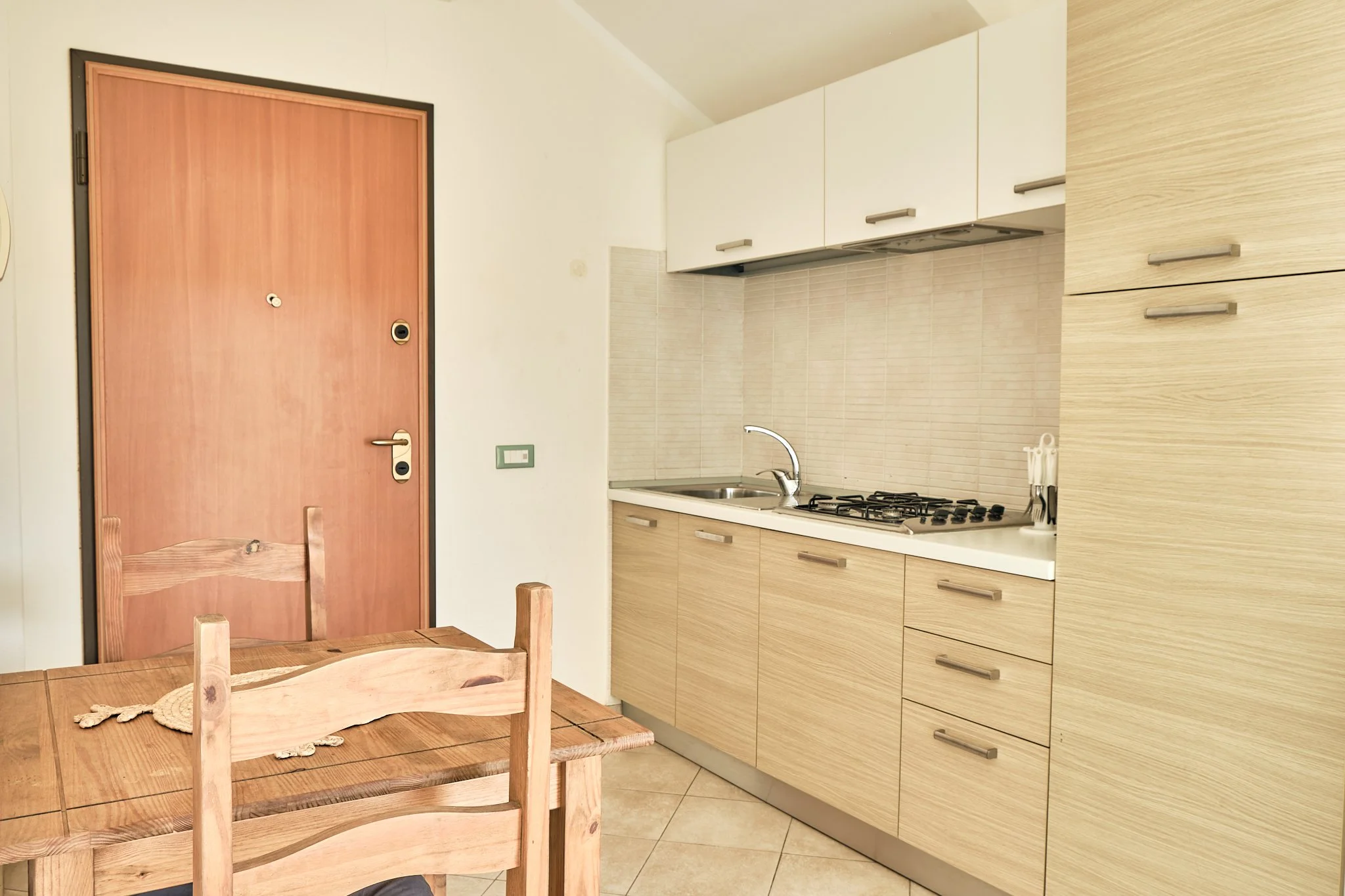 Kitchen with wooden dining table and chairs, cream-colored cabinets, beige tiled backsplash, stainless steel sink, gas stove, beige tiled floor, and wooden door.