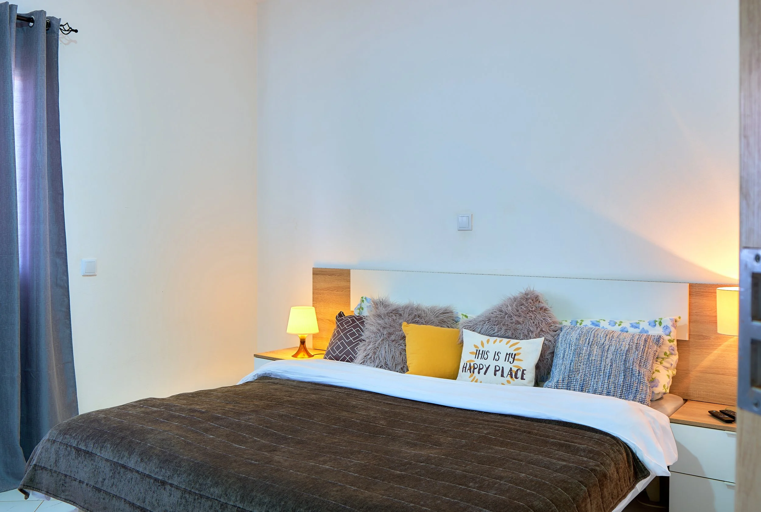 A bedroom with a bed covered in a dark brown quilt, decorated with various pillows including a white pillow with text and a fluffy gray one. There is a small wooden nightstand with a yellow lamp on it, and a beige wall with a light switch. To the lef