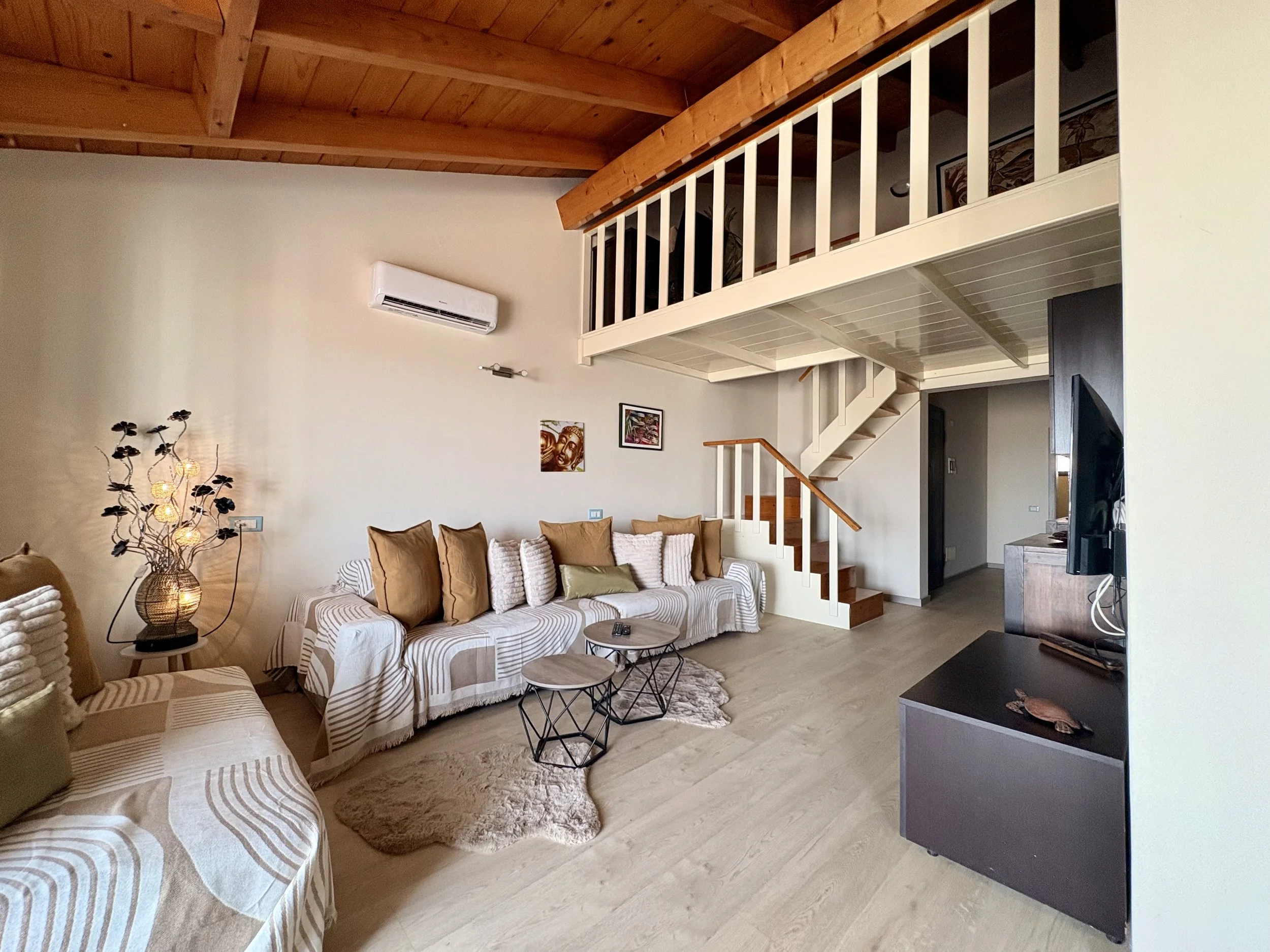 Living room with beige and white striped sofas, a wooden coffee table, a decorative lamp with flowers, artwork on the wall, a staircase, and a split air conditioning unit on the wall.