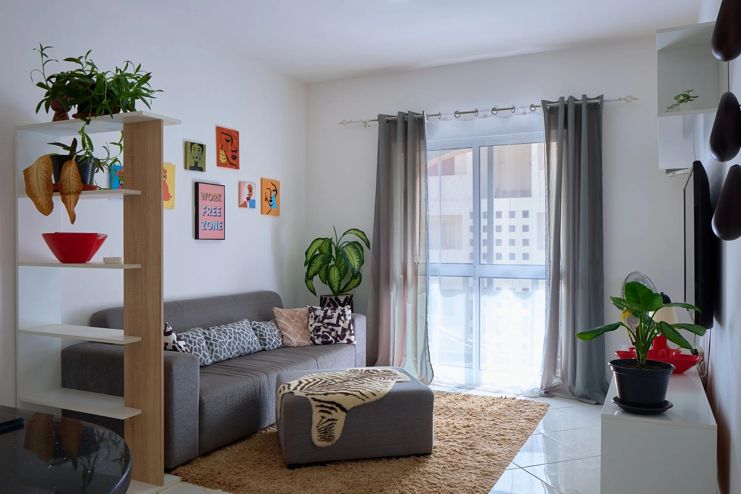 A cozy living room with a gray sofa, patterned pillows, a beige and black animal-print blanket, and a wooden and white shelving unit with plants. Wall art includes colorful portraits and a sign that says 'Work Free Zone.' There are large windows with