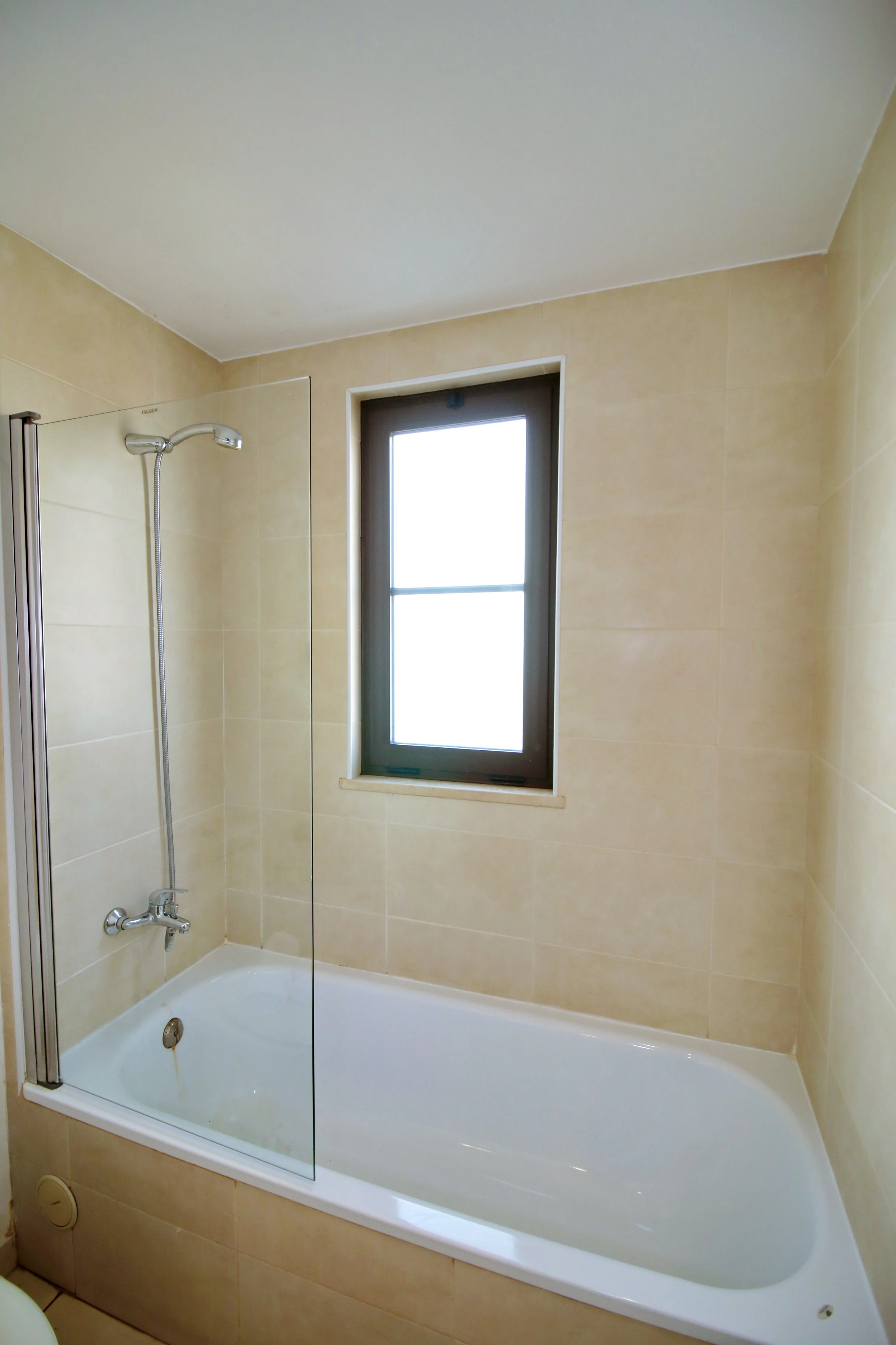 Bathroom bathtub with corner shower, a window above the tub, beige tiled walls, and a clear glass shower screen.