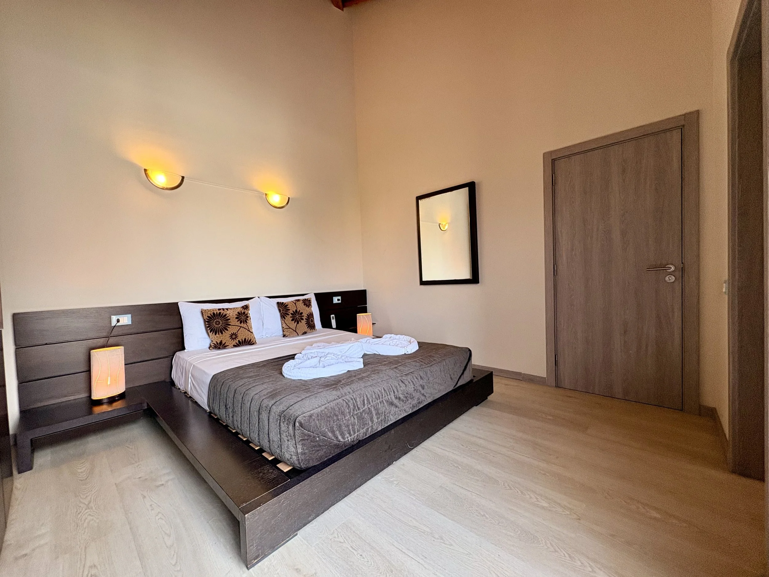 A bedroom with a bed, pillows, and bedside lamps, with a mirror on the wall and a closed wooden door.