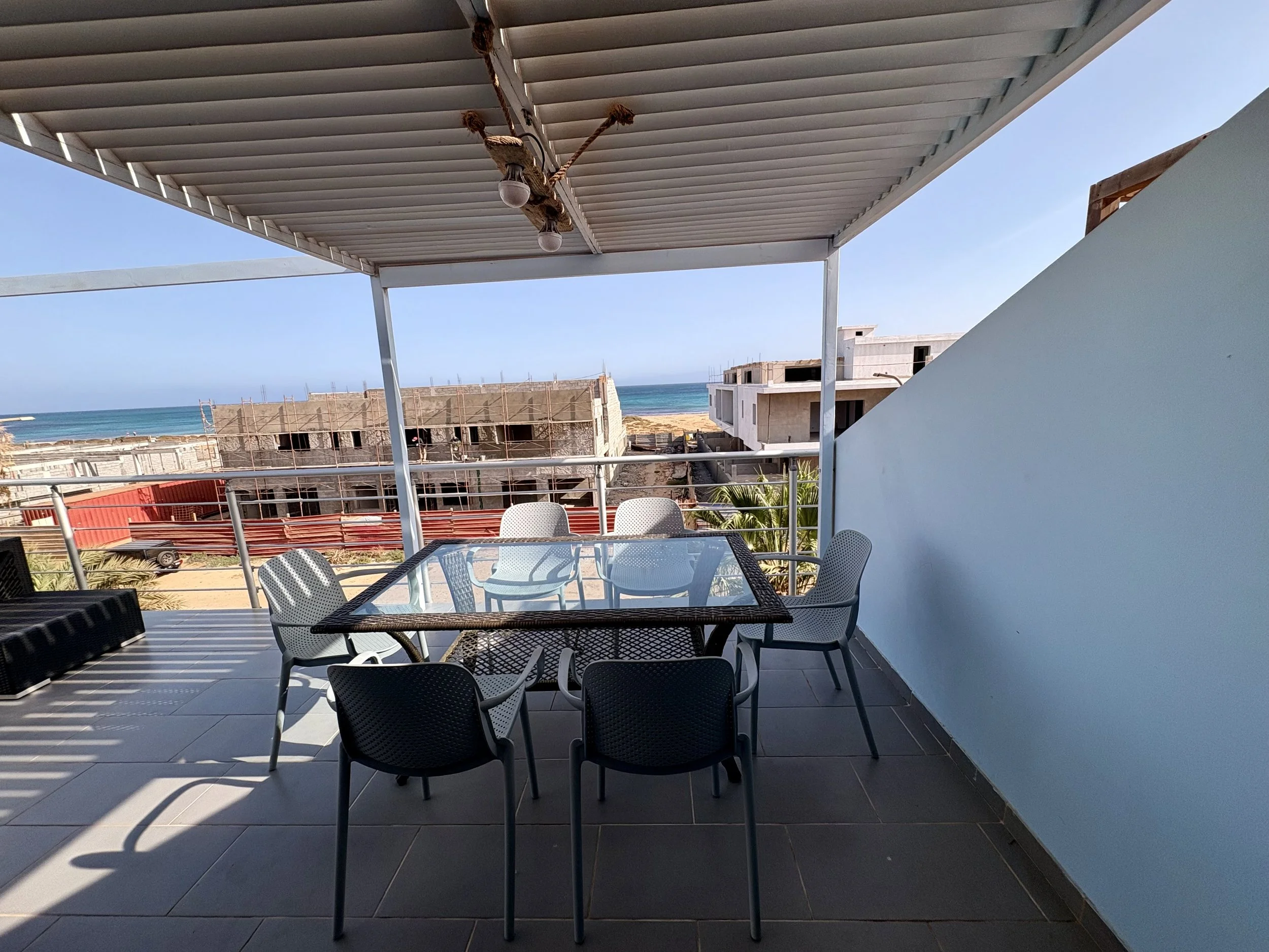 A balcony with a table and multiple chairs, overlooking a beach with buildings under construction, blue ocean and sky in the background, and a white wall on the right.