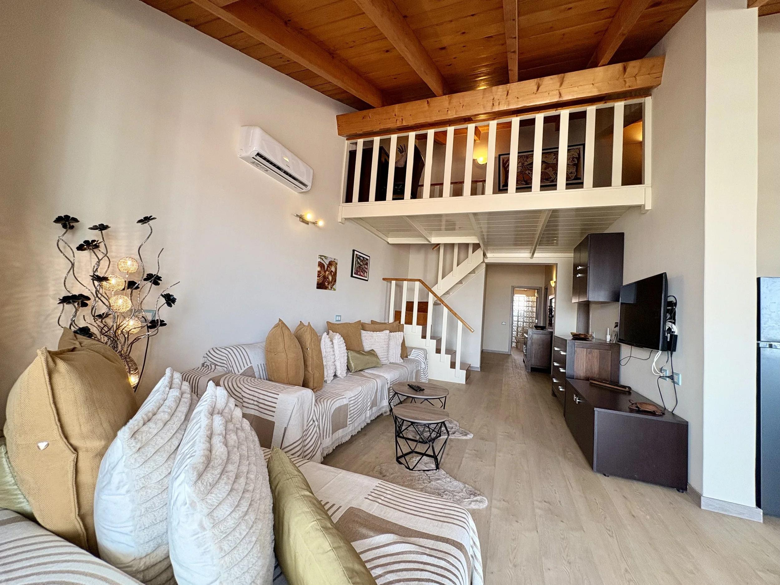 Living room with beige sofa, pillows, wooden ceiling, staircase, wall decorations, TV, and kitchen area.