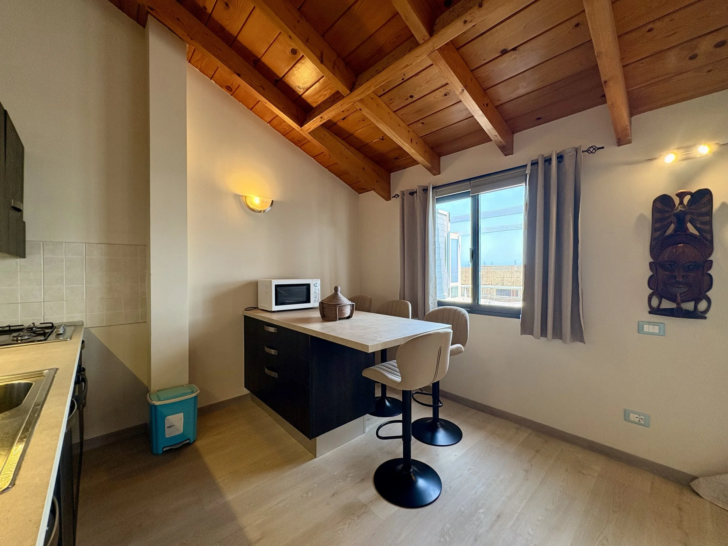 A kitchen with a wooden ceiling, white walls, a window with curtains, a small table with chairs, a microwave, and tribal wall art.