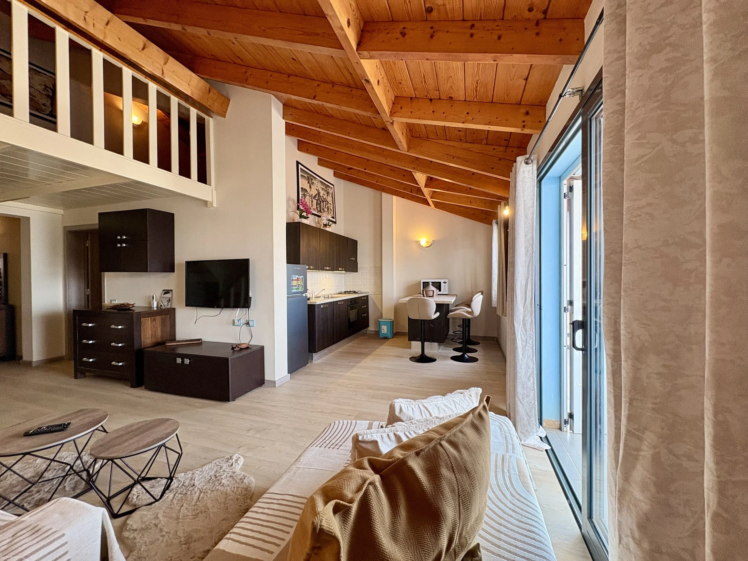 Living room with wooden ceiling, open kitchen with dark cabinets, small dining area, couch with pillows, sliding glass door with curtains.