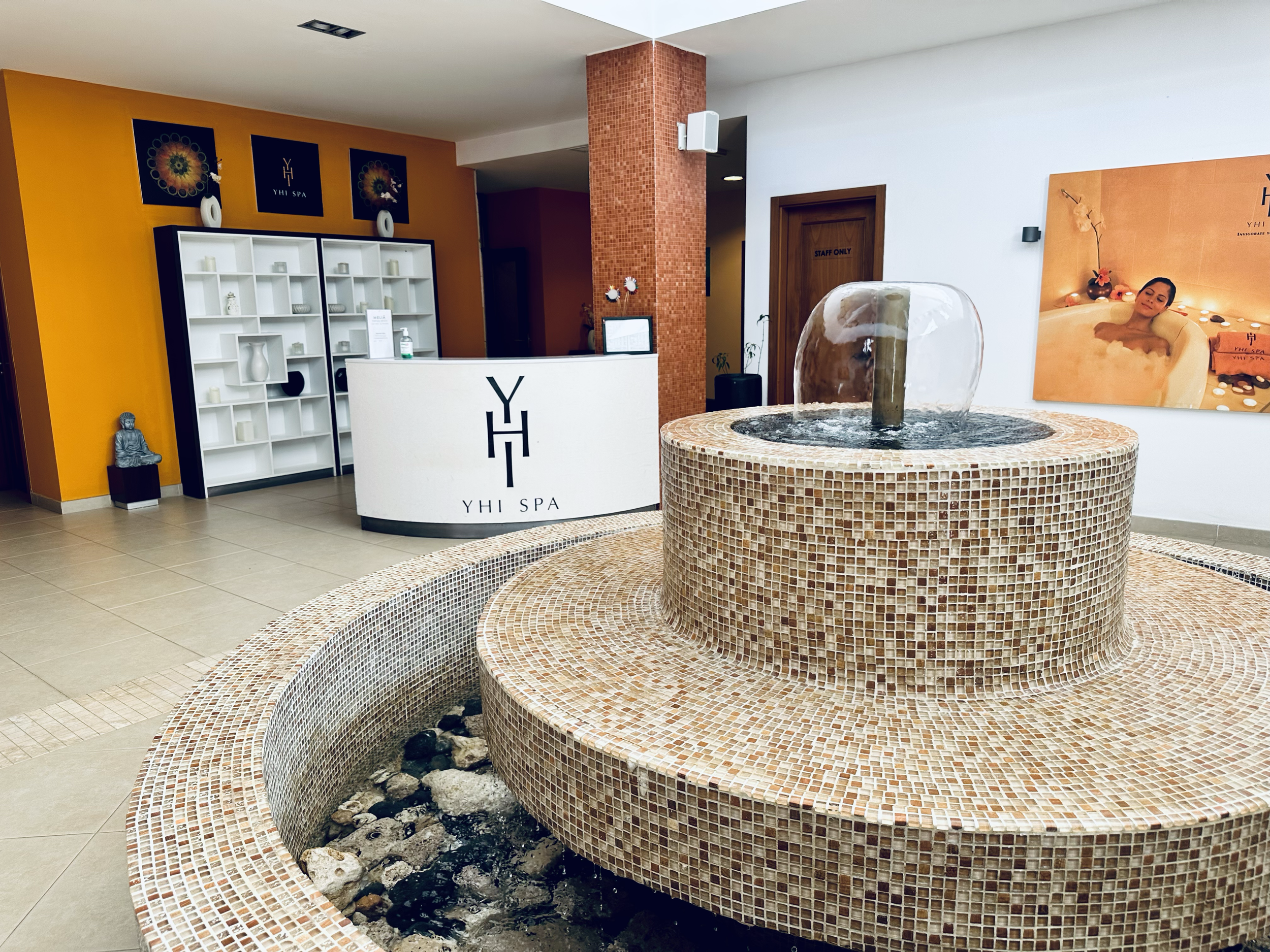 Spa reception area with a fountain, white reception desk with YHI SPA logo, orange and white walls, tropical decor, and a large photograph of a woman relaxing in a bath with candles and flowers.