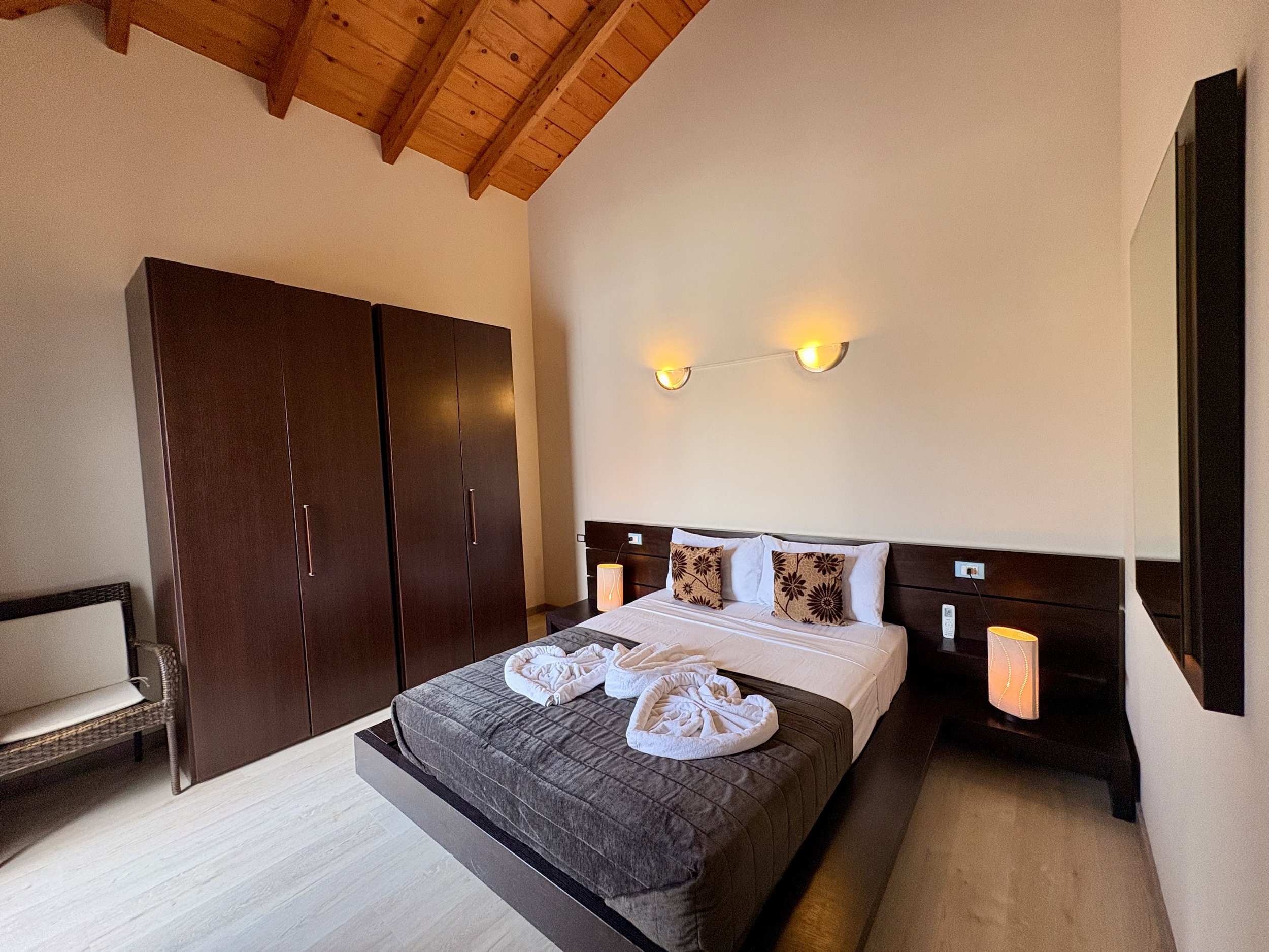 A bedroom with a sloped wooden ceiling, dark wooden wardrobe, bed with pillows, bedside lamps, and a small chair.