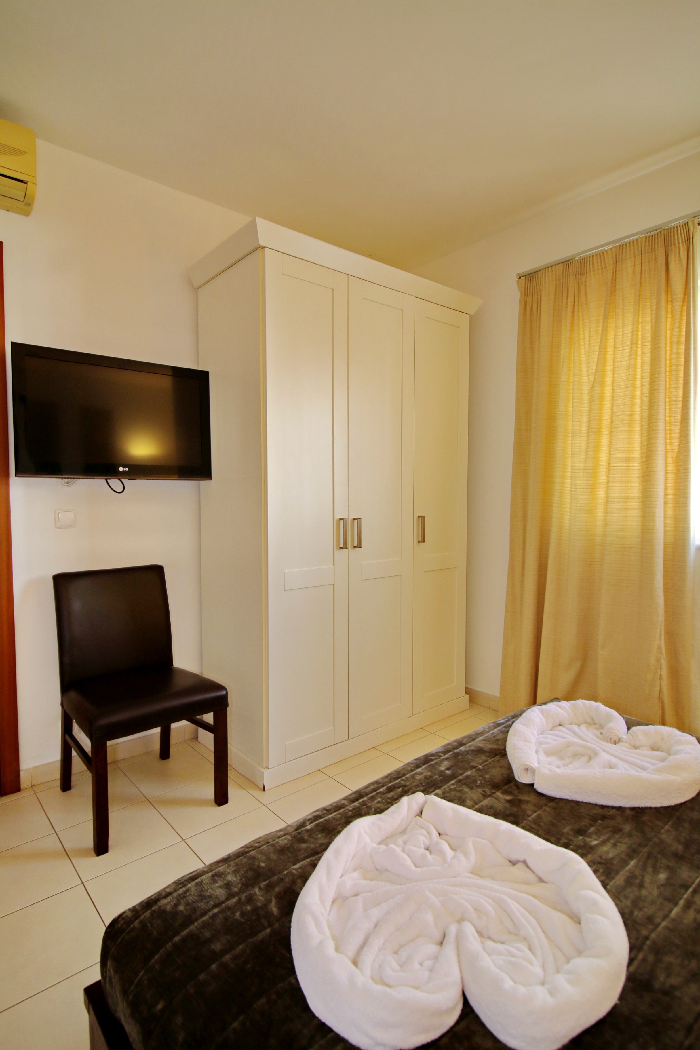 Hotel room with a bed, two towl swans, a white wardrobe, a chair, a wall-mounted TV, a curtain, and an air conditioning unit.
