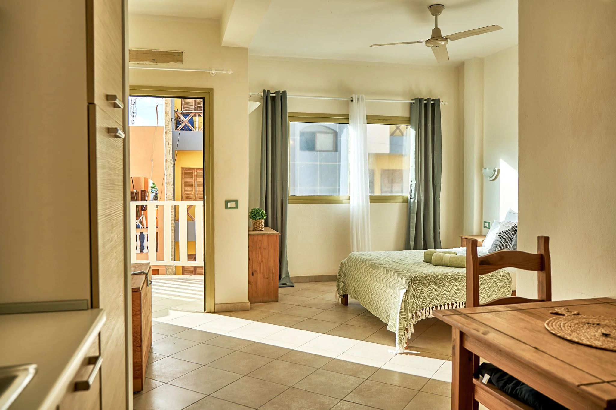 Interior of a bright hotel room with a bed, small wooden side table, large windows with curtains, ceiling fan, and a balcony door opening to an outdoor area.