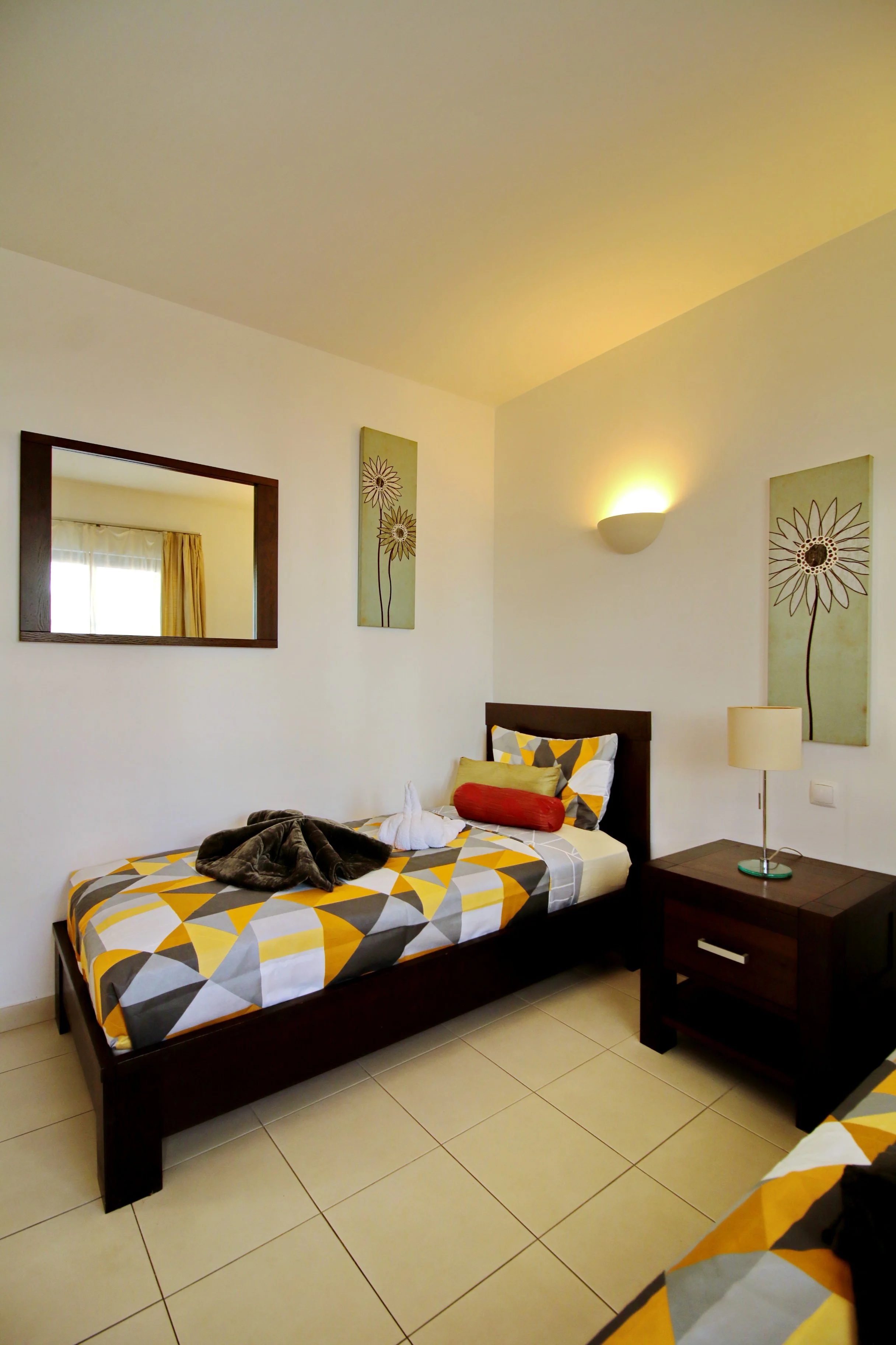 A bedroom with a single bed, a pillow, a blanket, and a towel folded into a swan shape on top. There are framed flower artworks on the wall and a mirror, a wall-mounted light, a nightstand with a lamp, and tiled flooring.