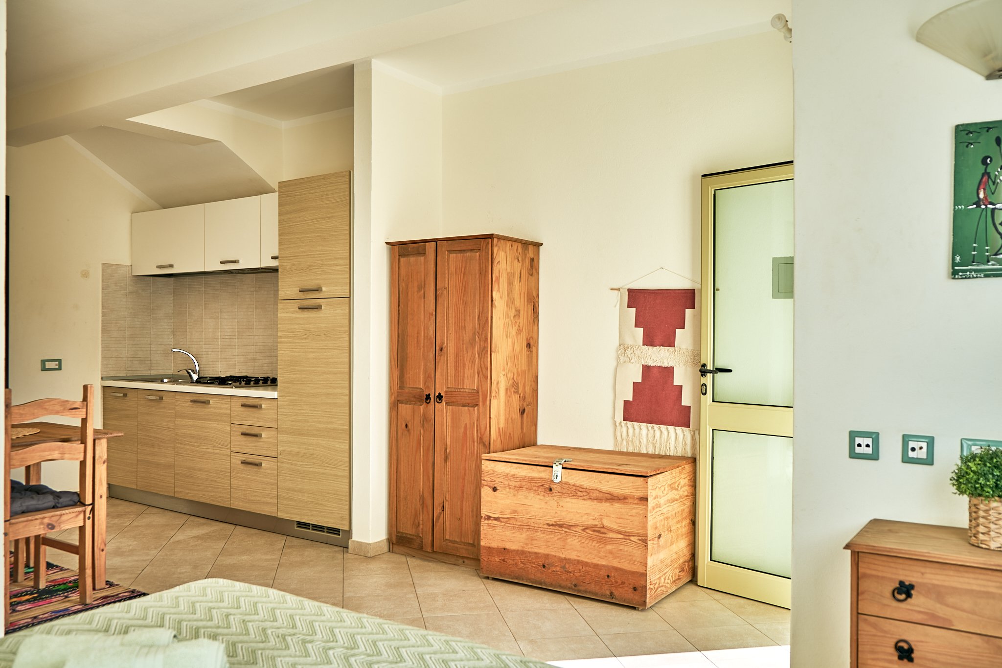 Interior view of a room with a kitchenette on the left, including wooden cabinets, a sink, and a stove. There is a wooden storage chest and a small wooden table with a drawer in the center, and a white door on the right. The room has a modern, cozy d
