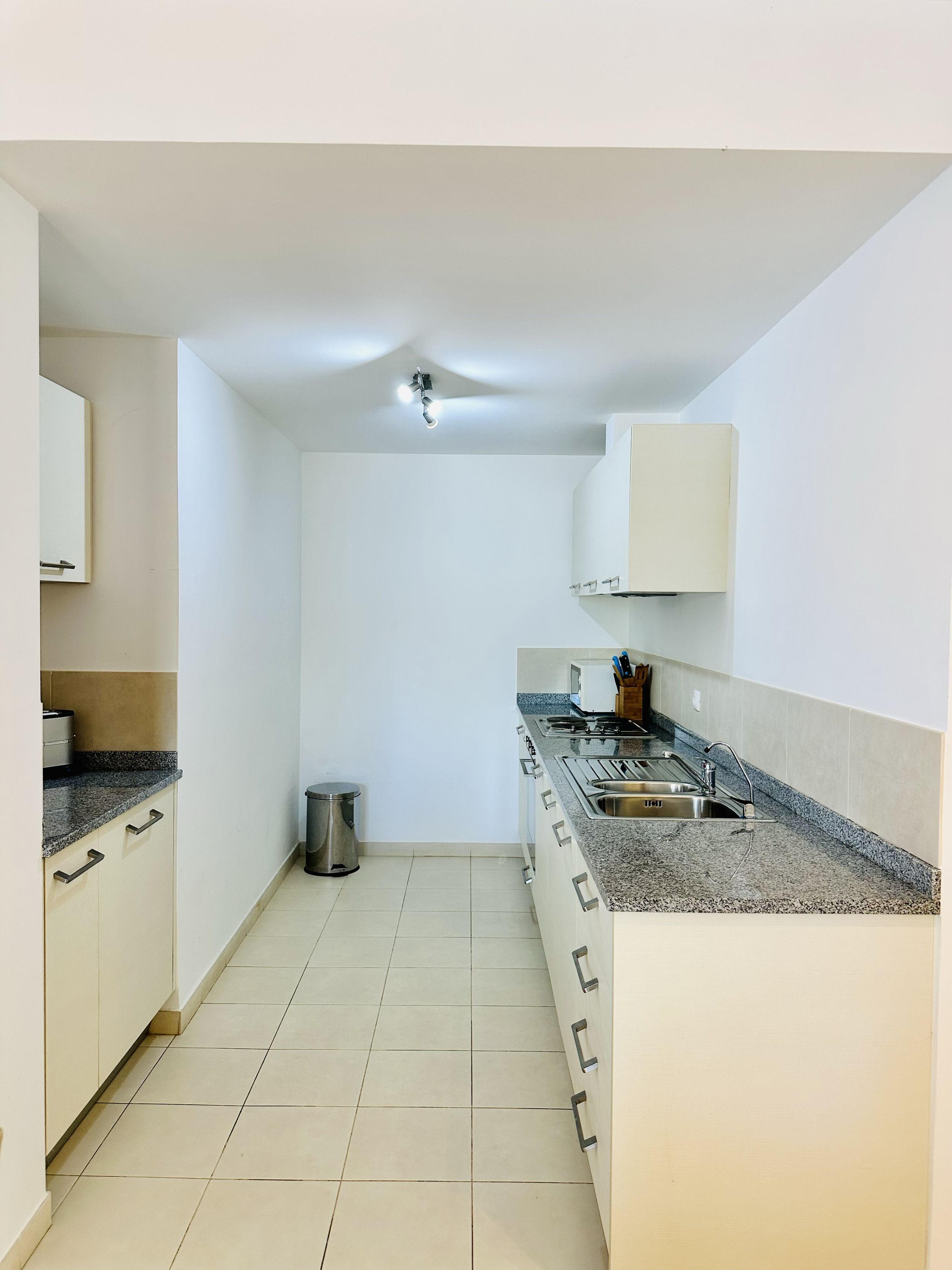 A clean, modern kitchen with white cabinets, granite countertops, a stainless steel sink, a stove, and a microwave. There are white tiled floors and walls with minimal decor.