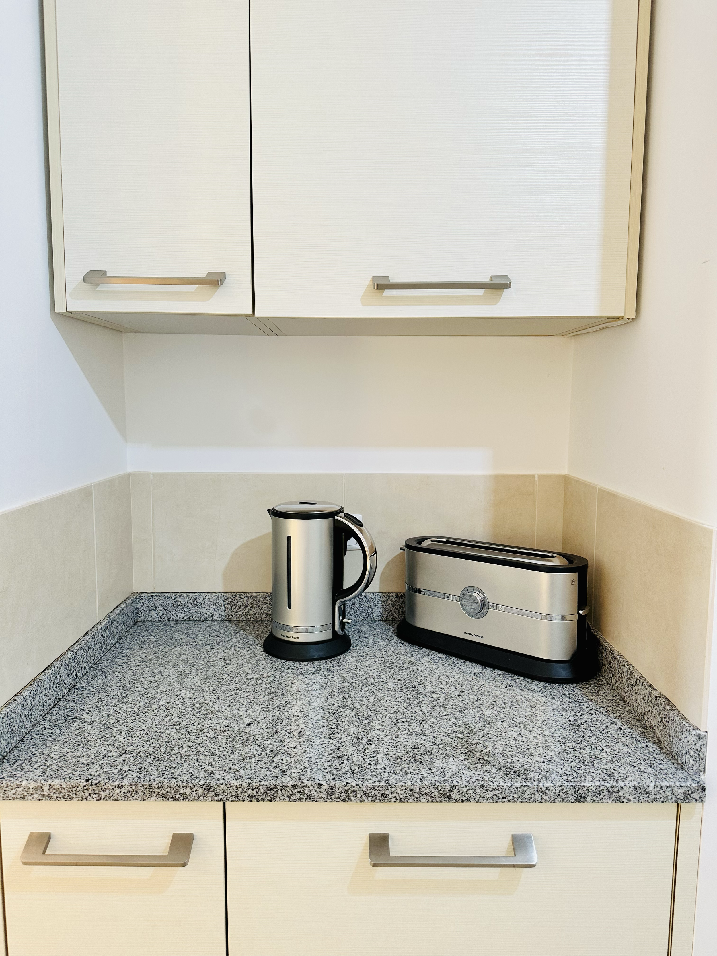 Kitchen counter with a silver electric kettle and a silver toaster, above white cabinets with metallic handles.