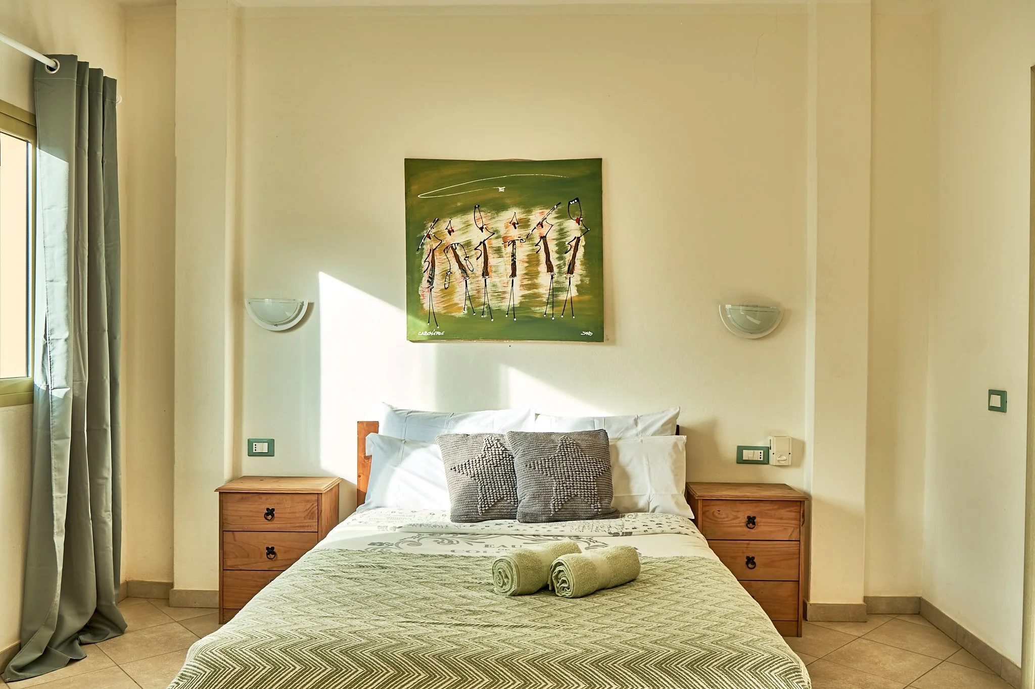 A bedroom with a bed covered in a green patterned bedspread and two rolled towels on top. There are two wooden nightstands on either side of the bed, each with a drawer and a round black handle. The wall behind the bed has a painting of abstract figu