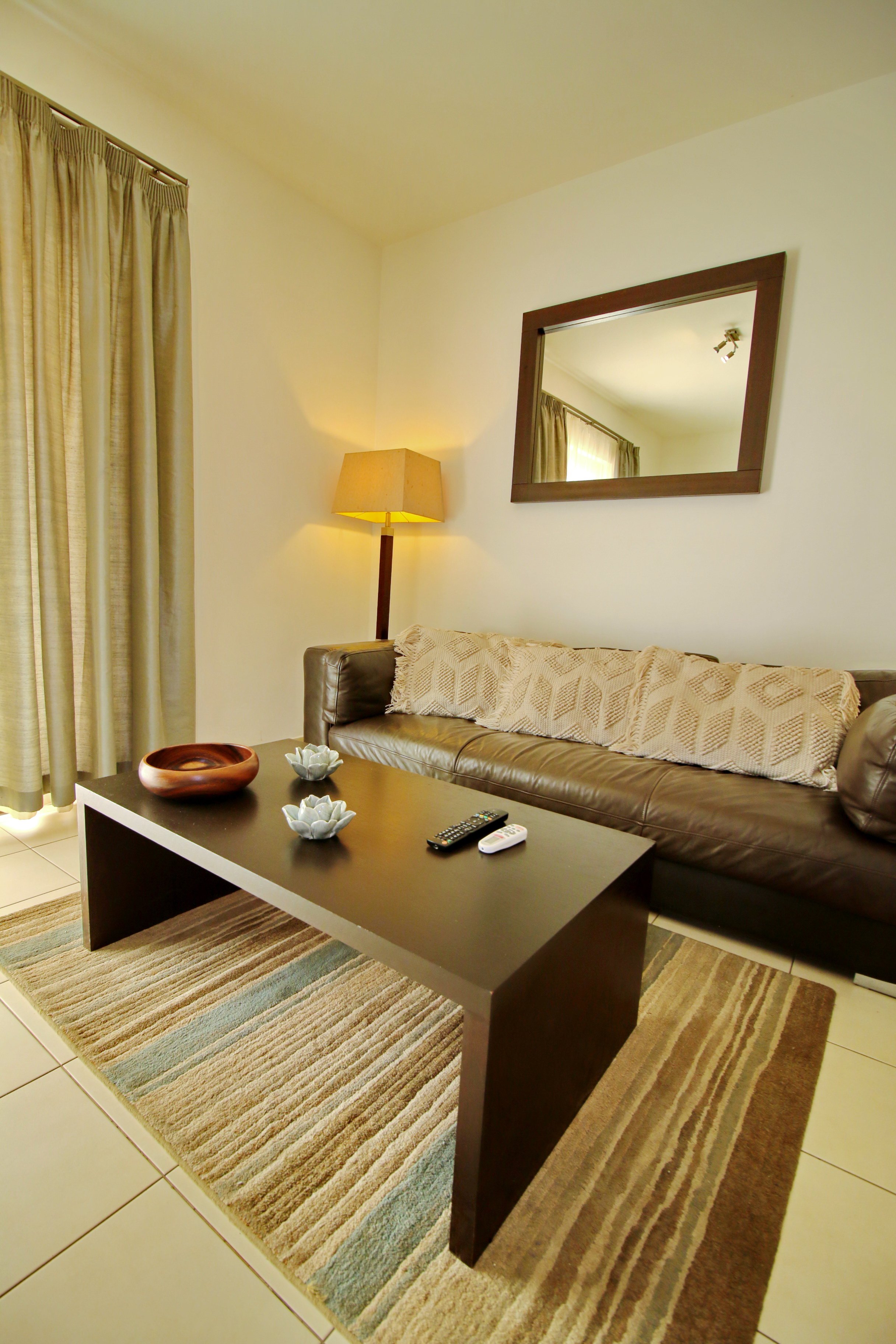 Living room with a striped rug, beige curtains, a beige sofa with a decorative blanket, a wooden coffee table with bowls and remote controls, a floor lamp, and a mirror on the wall reflecting the curtains and ceiling lights.