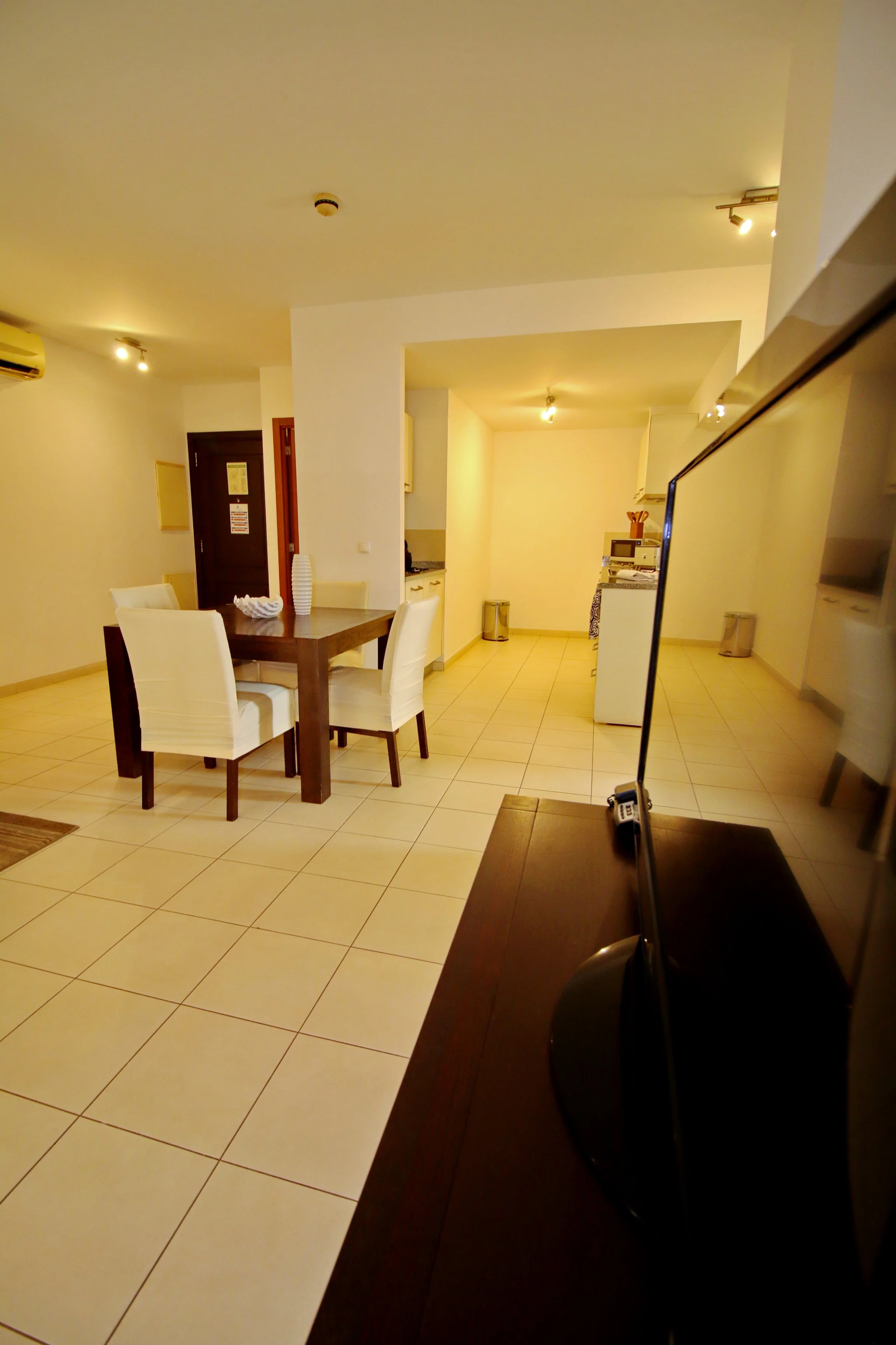 Living room with dining table, white chairs, kitchen appliances, and a TV, illuminated by ceiling lights and wall-mounted lamps.